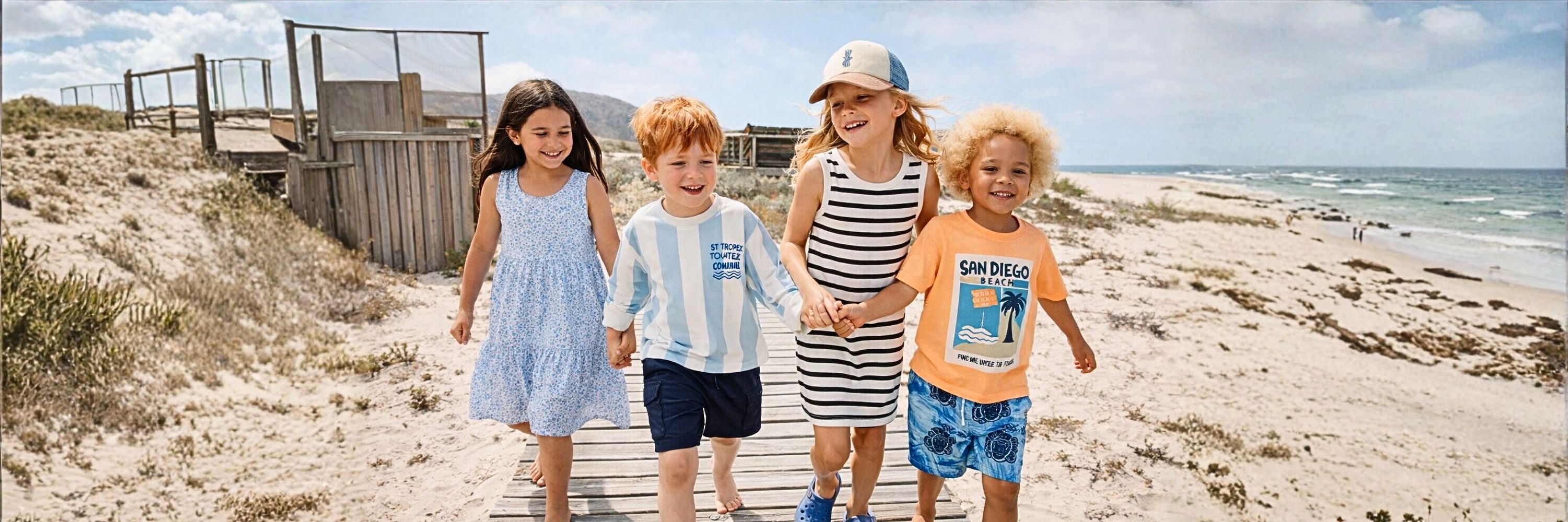 Four children walking together on a wooden path by the beach, smiling and holding hands, wearing casual summer outfits with the sea and sandy shore in the background.