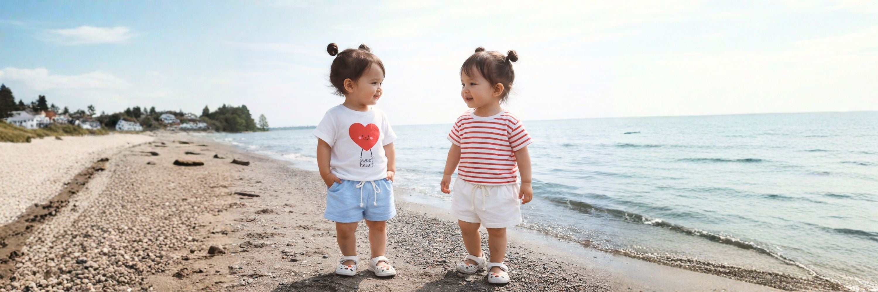 Two toddlers stand on a beach facing each other, smiling in casual summer outfits as gentle waves roll in beside them.