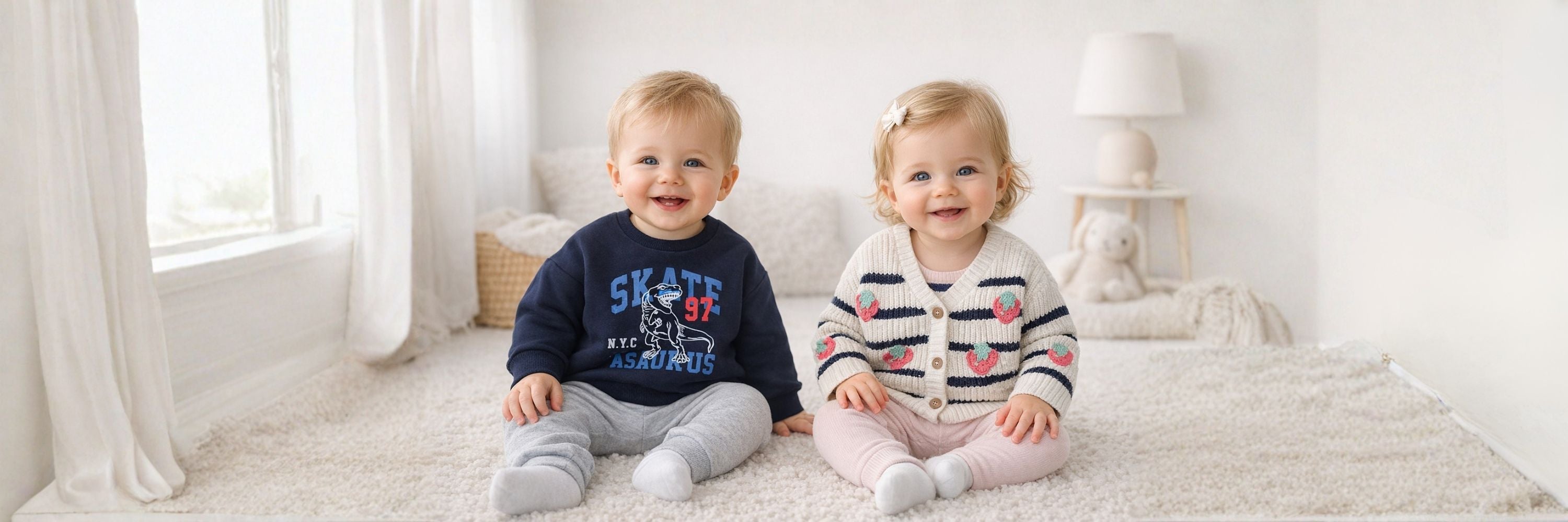Two smiling babies sitting on a soft carpet indoors, wearing cozy outfits, with a bright window and neutral home setting in the background.