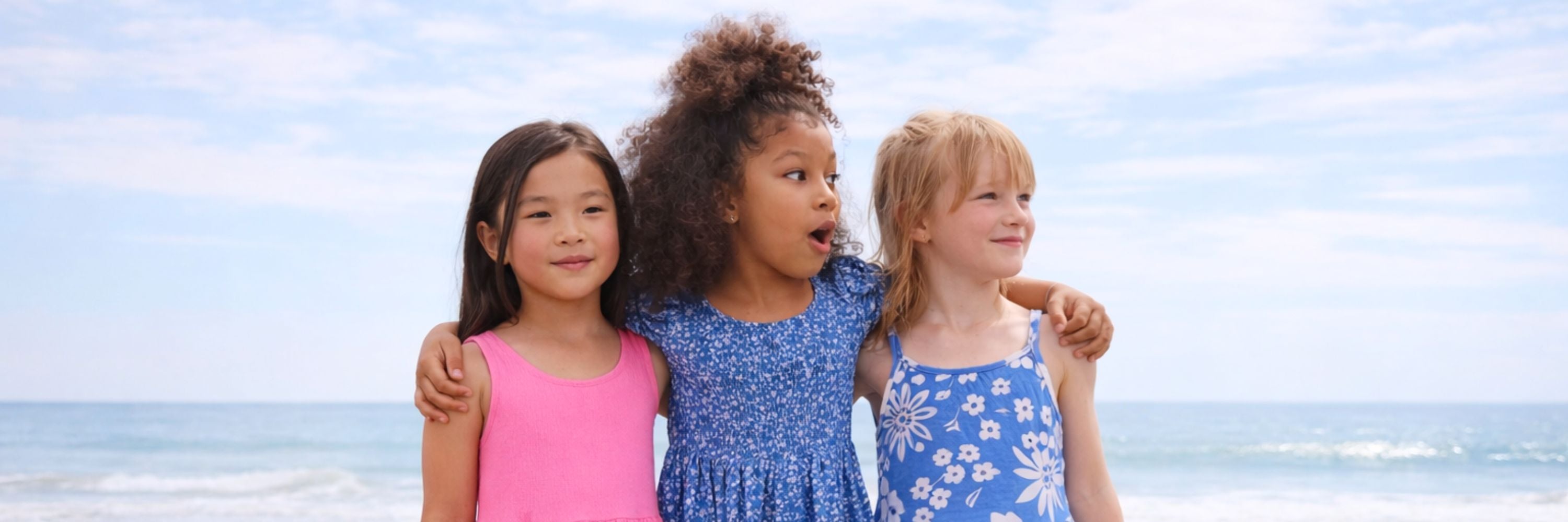 Three girls standing together at the beach with their arms around each other, smiling and looking away, wearing colorful summer dresses with the ocean in the background.