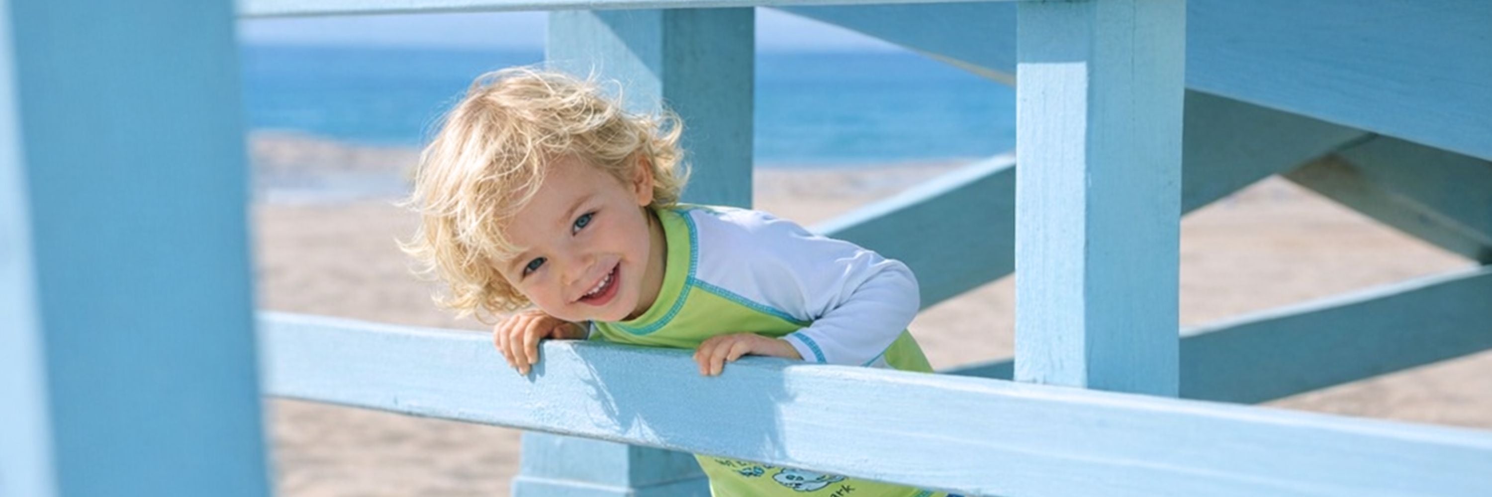 A smiling toddler leaning on a wooden beach railing, wearing a long-sleeve swim top, with the ocean and sandy beach in the background.