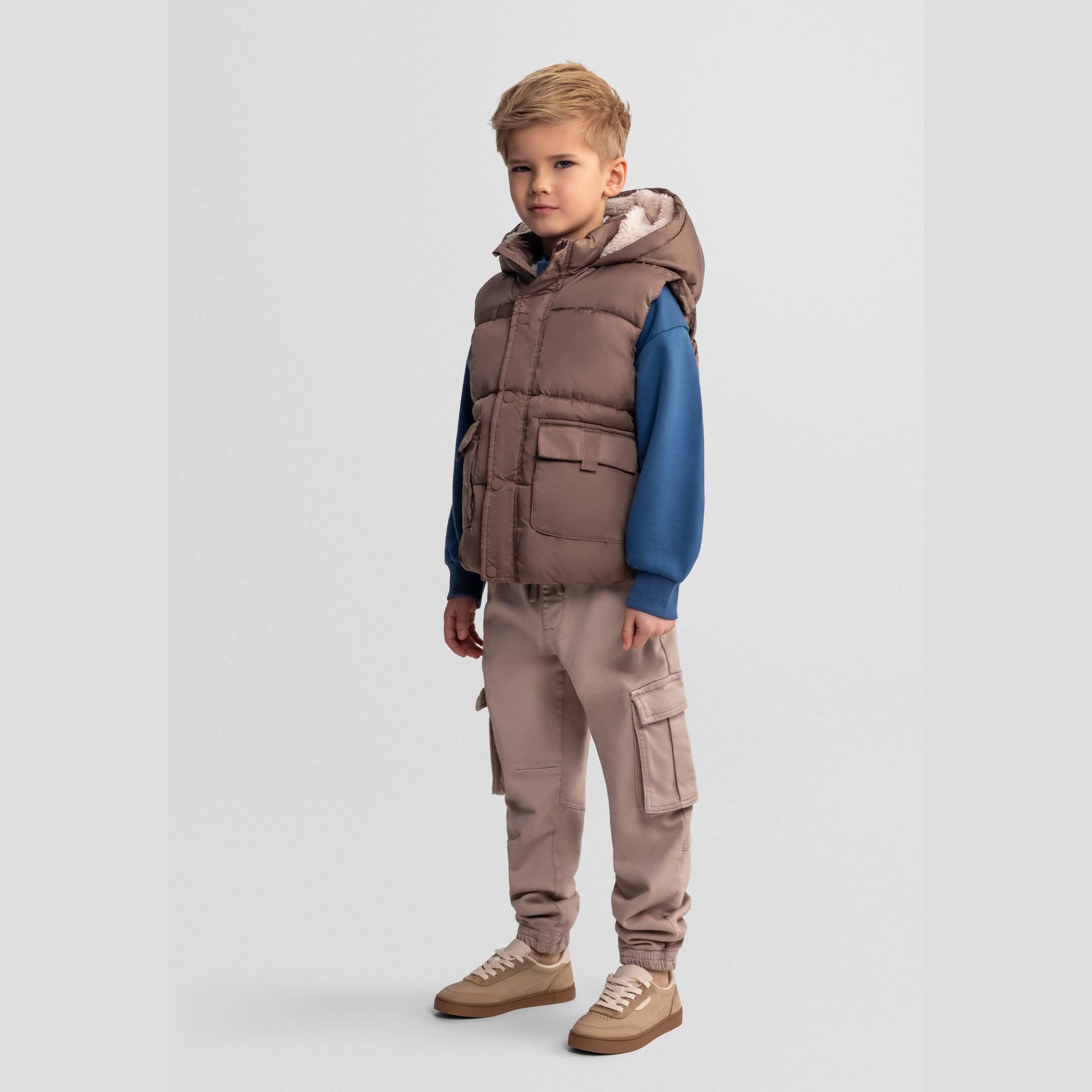 Front view of boy wearing beige quilted hooded gilet over blue sweatshirt with beige cargo trousers on grey background