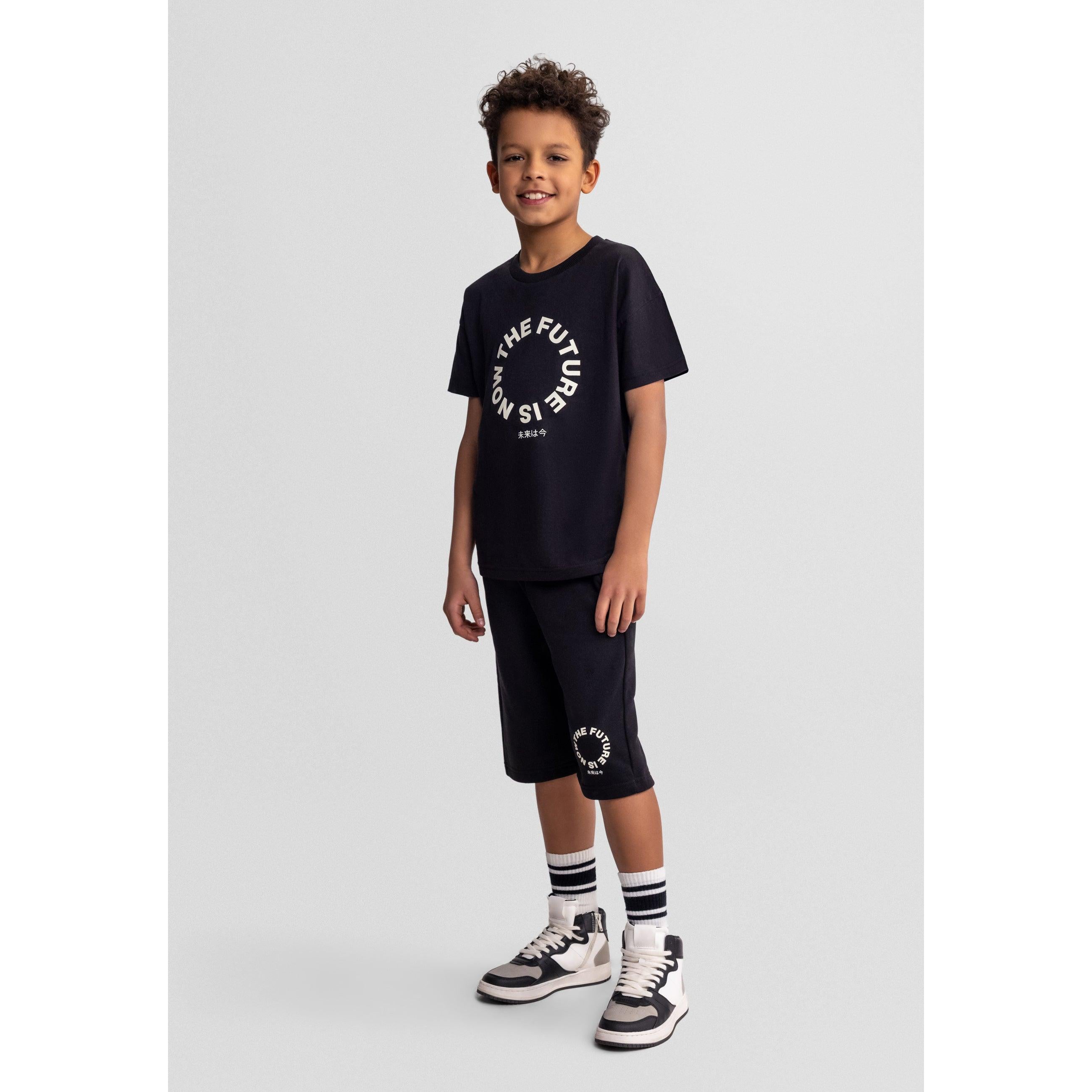 Young boy wearing black t-shirt and shorts set with The Future is Now circular print, standing against grey background wearing striped socks and trainers