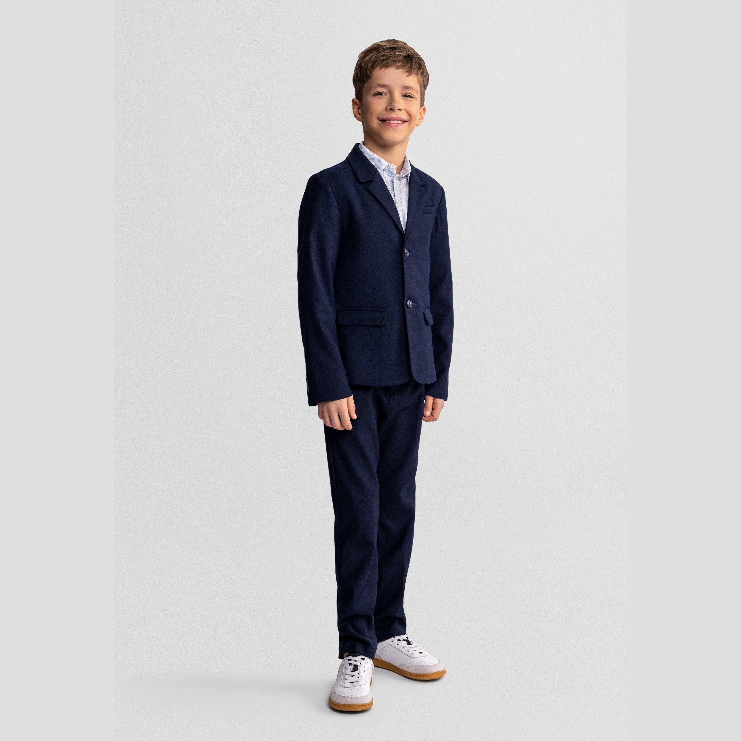 Young boy with curly hair wearing complete navy suit with tie, standing formally against white studio background