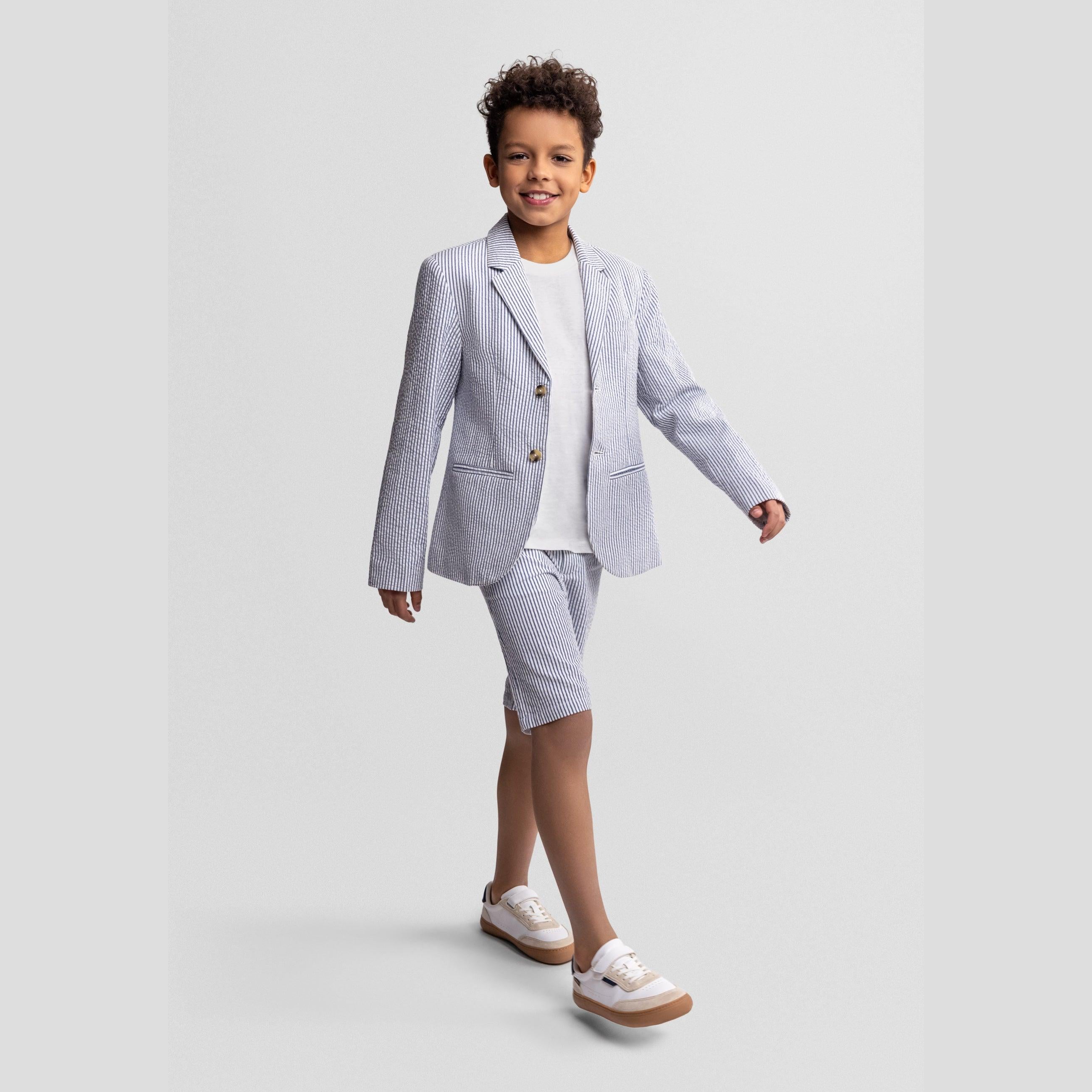 Boy in navy striped blazer and matching shorts with white top, posing with one hand on hip against light grey studio background
