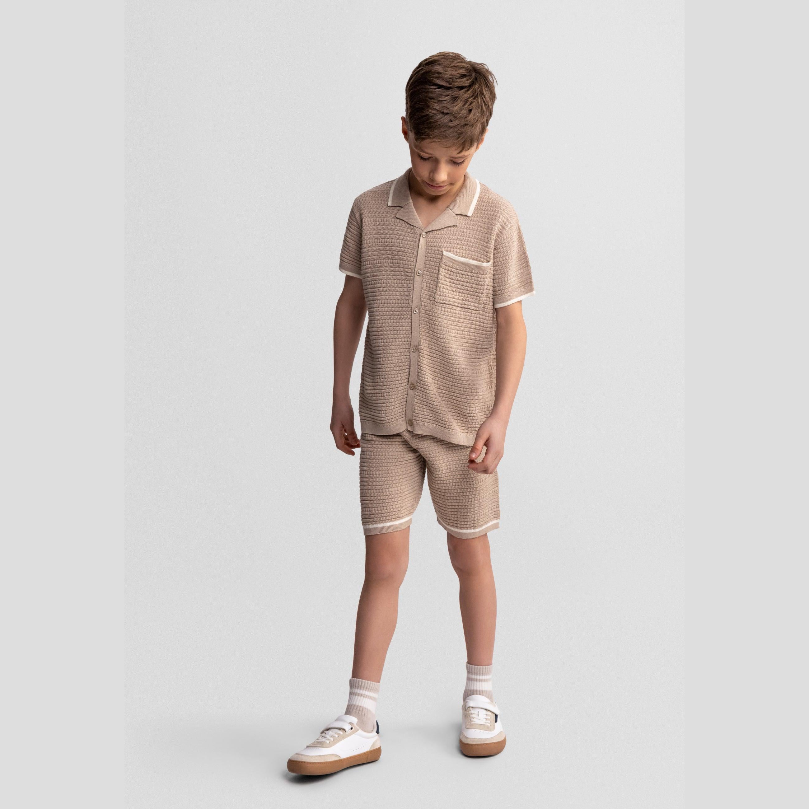 Boy wearing beige knitted polo shirt and matching shorts set against light grey background, styled with white sneakers and socks