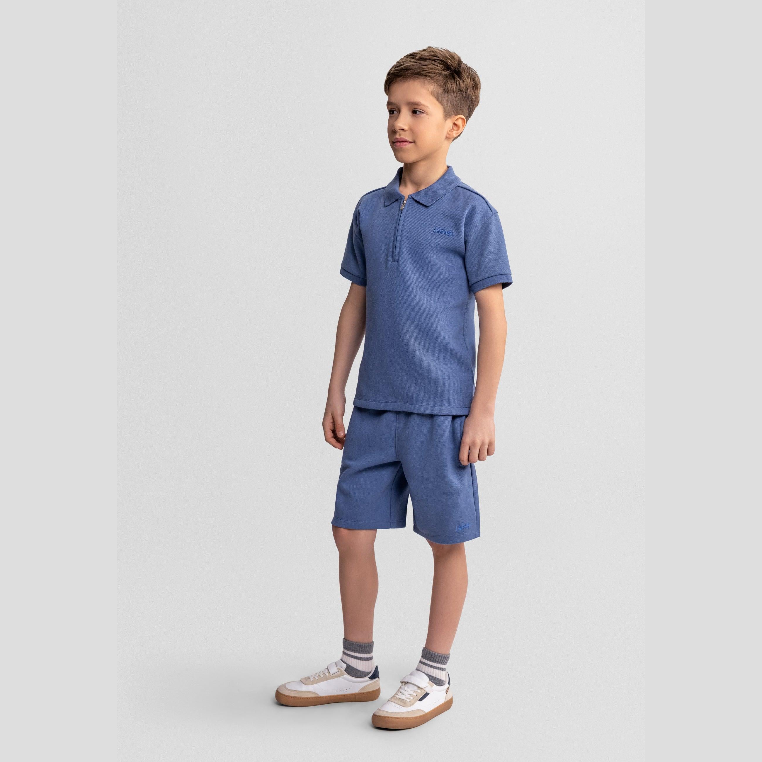 Young boy wearing blue pique polo shirt and matching shorts set, standing against white background, casual pose looking to the side