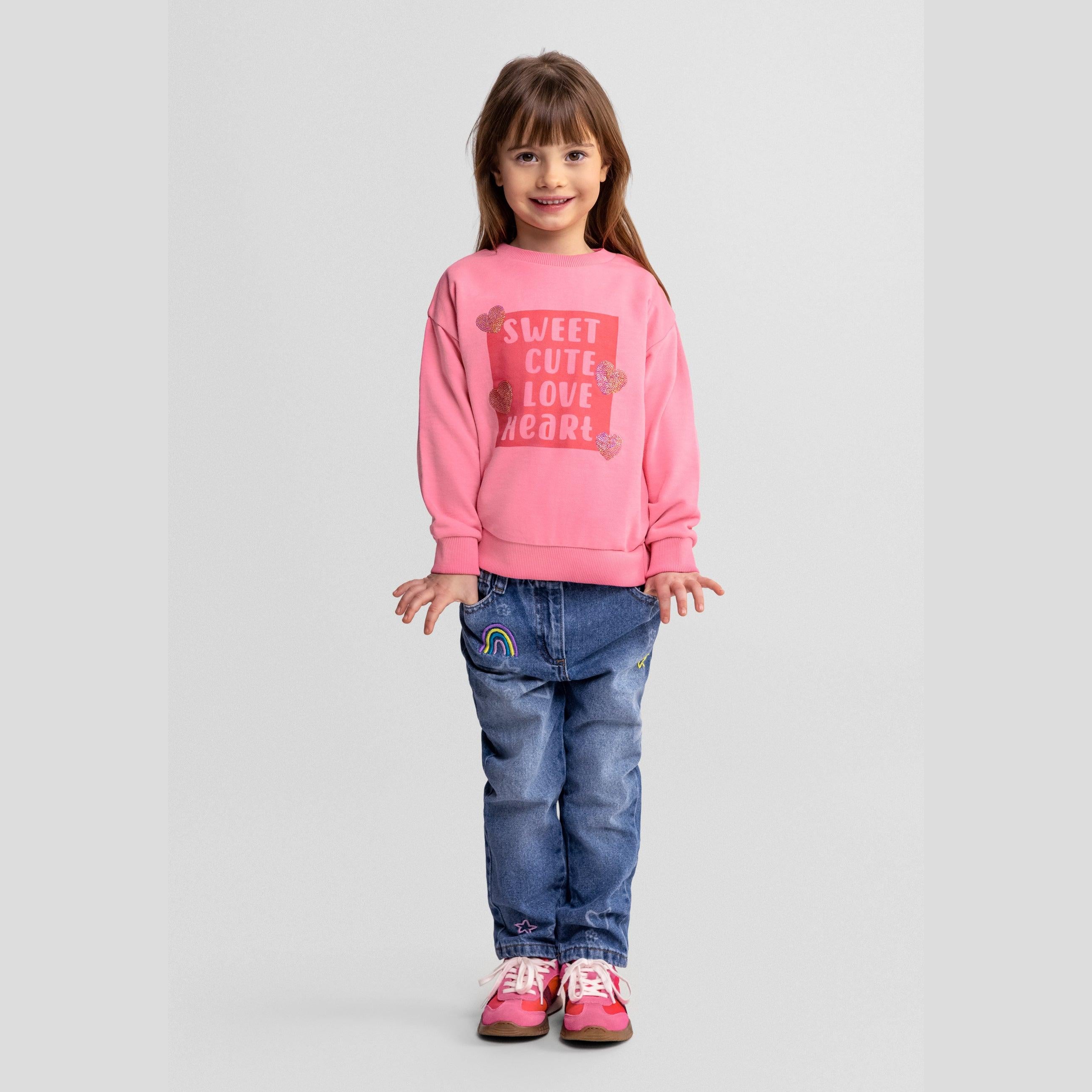 Young girl wearing pink drop shoulder sweatshirt with 'Sweet Cute Love Heart' print and sequined hearts, paired with denim jeans and pink sneakers, standing against light background