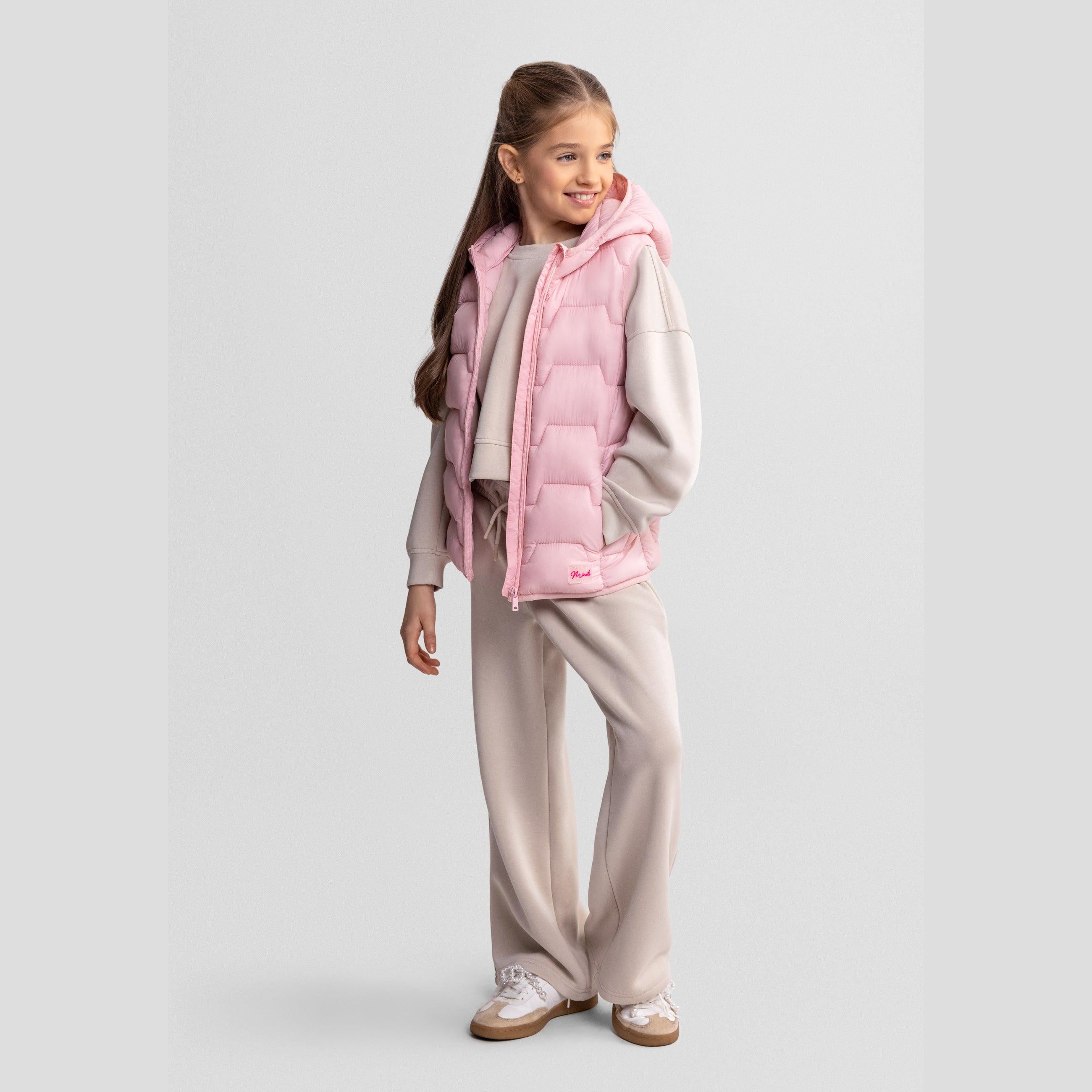 Young girl smiling wearing pink quilted gilet over beige tracksuit on light grey studio background