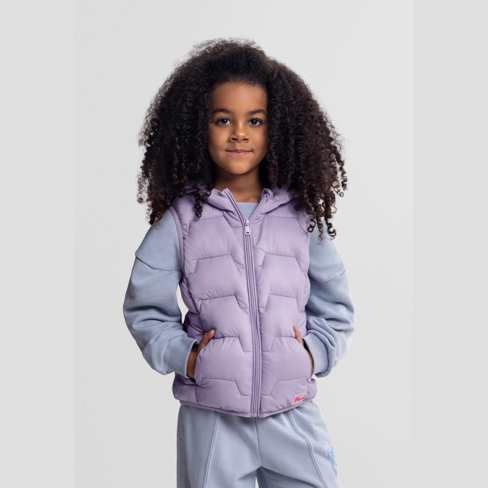 Young girl wearing lilac padded gilet with grey hoodie and tracksuit bottoms, smiling against light background