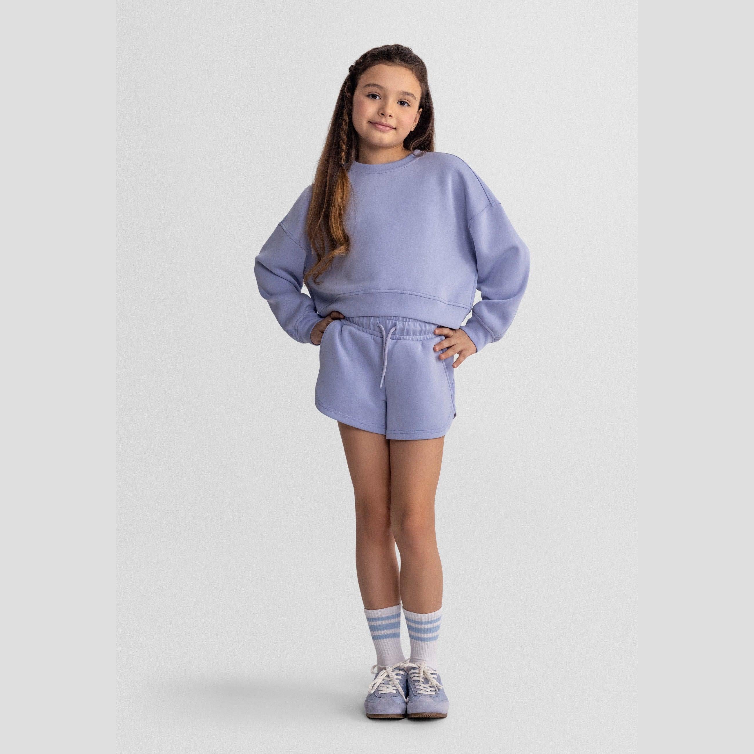 Young girl wearing lavender sweatshirt and shorts set with matching striped socks and sneakers on white background