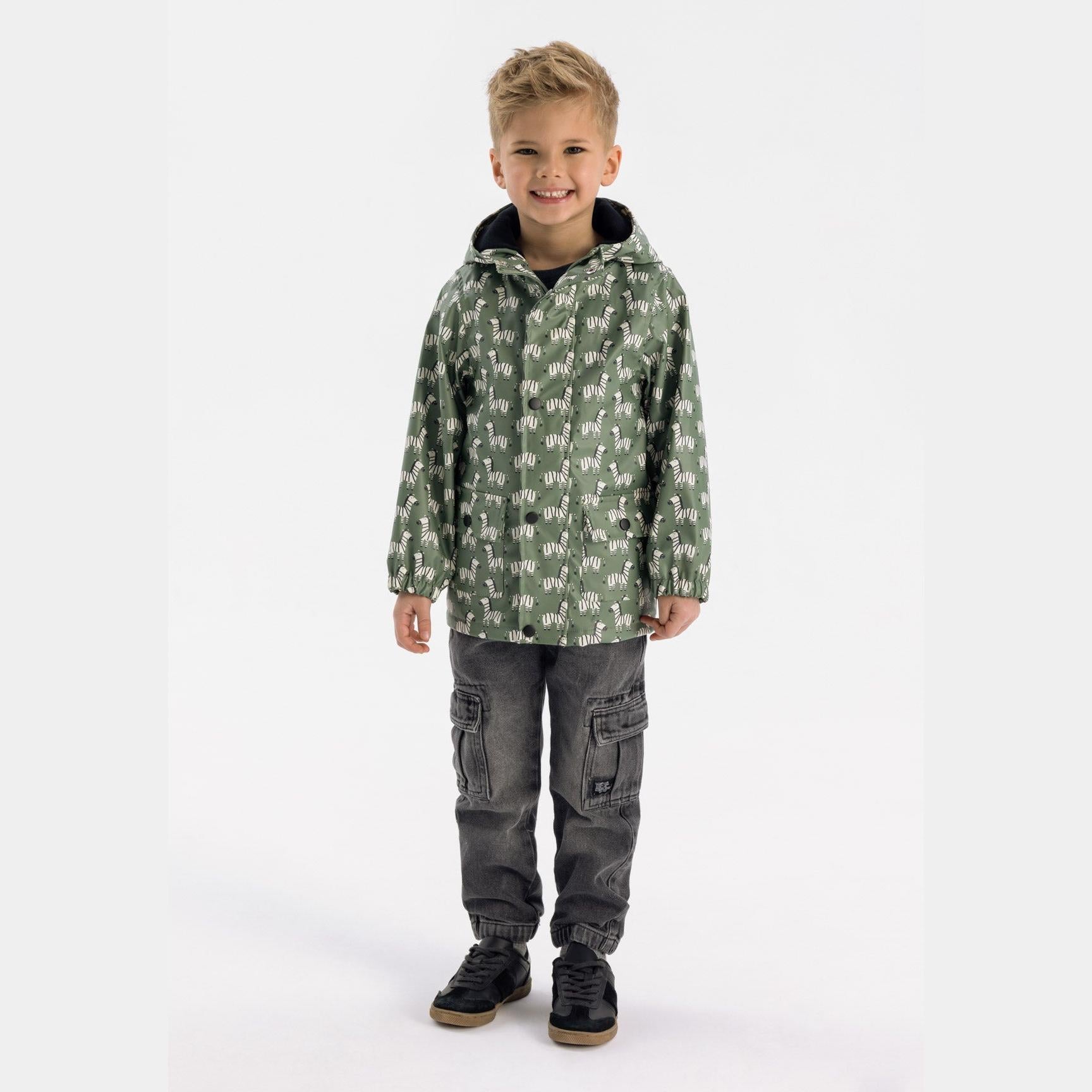 Young boy wearing khaki rain jacket with zebra print pattern and hood, paired with grey cargo trousers and black trainers, standing against white studio background