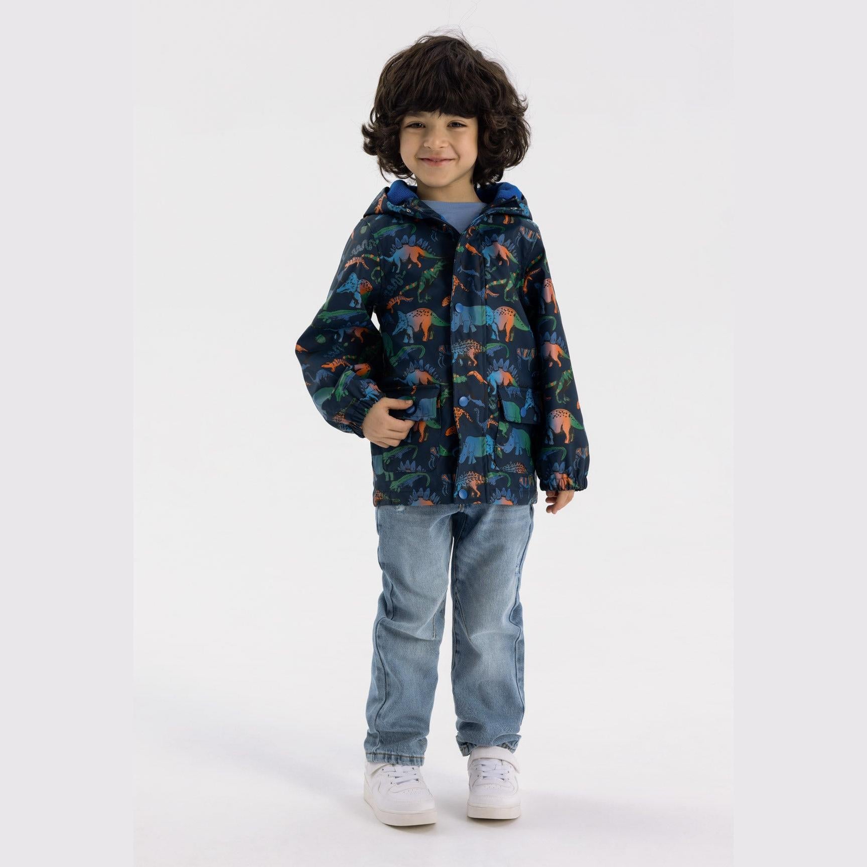 Young child wearing navy rain jacket with colourful dinosaur print, paired with light blue jeans and white trainers, smiling against white studio background