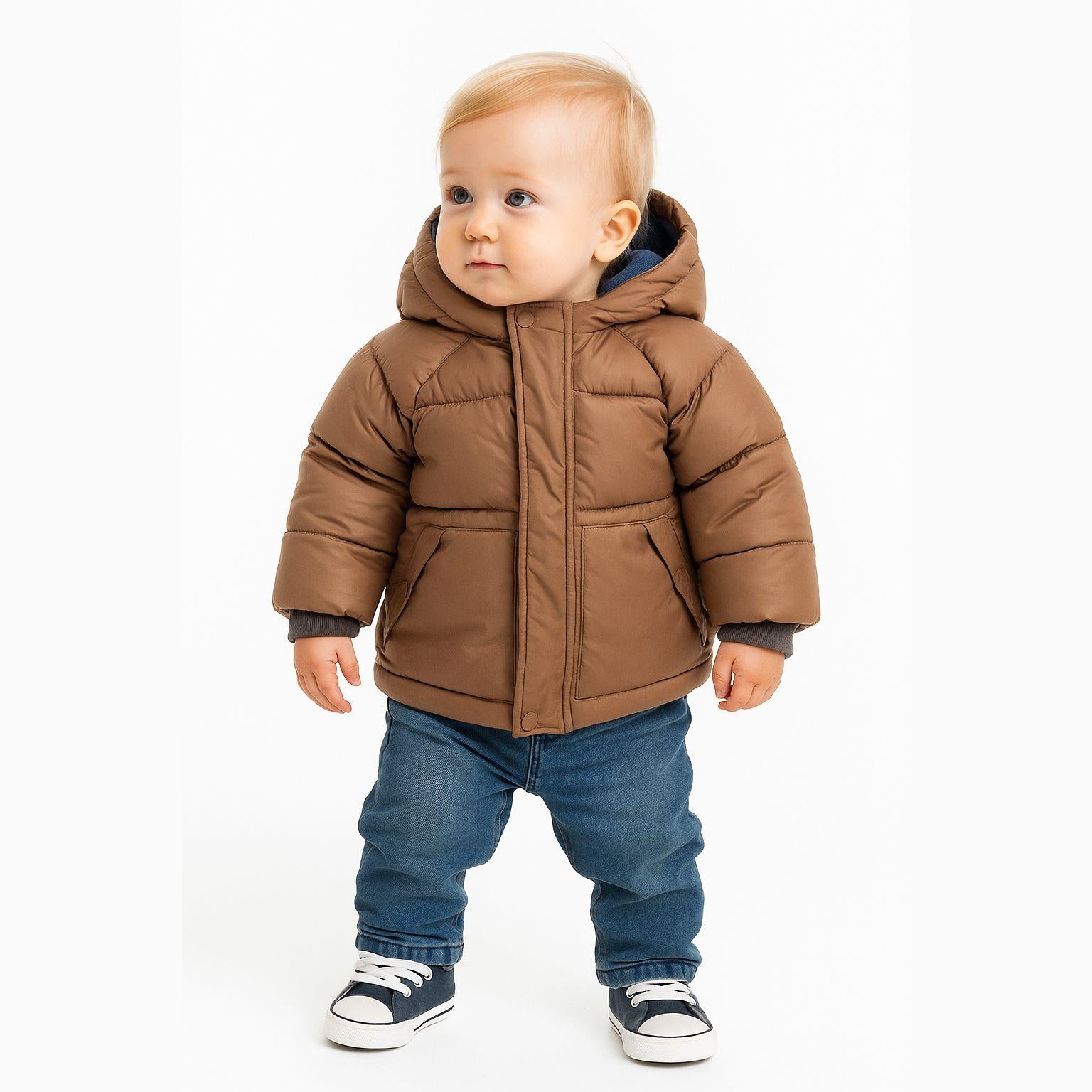Baby wearing brown quilted padded jacket with hood and blue jeans, standing against white background