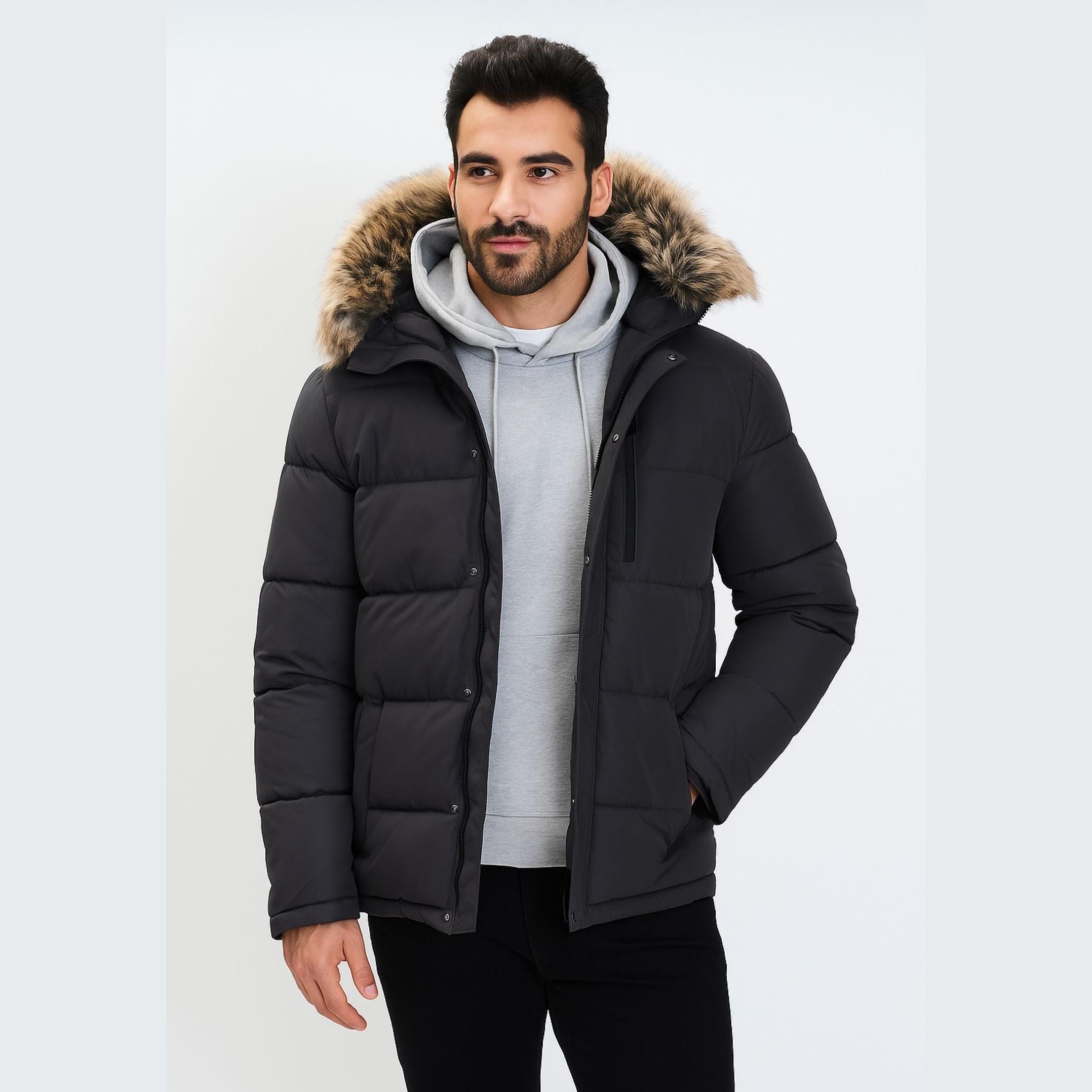 Adult male model wearing grey puffer jacket with faux fur hood over grey hoodie, hands in pockets, white studio background