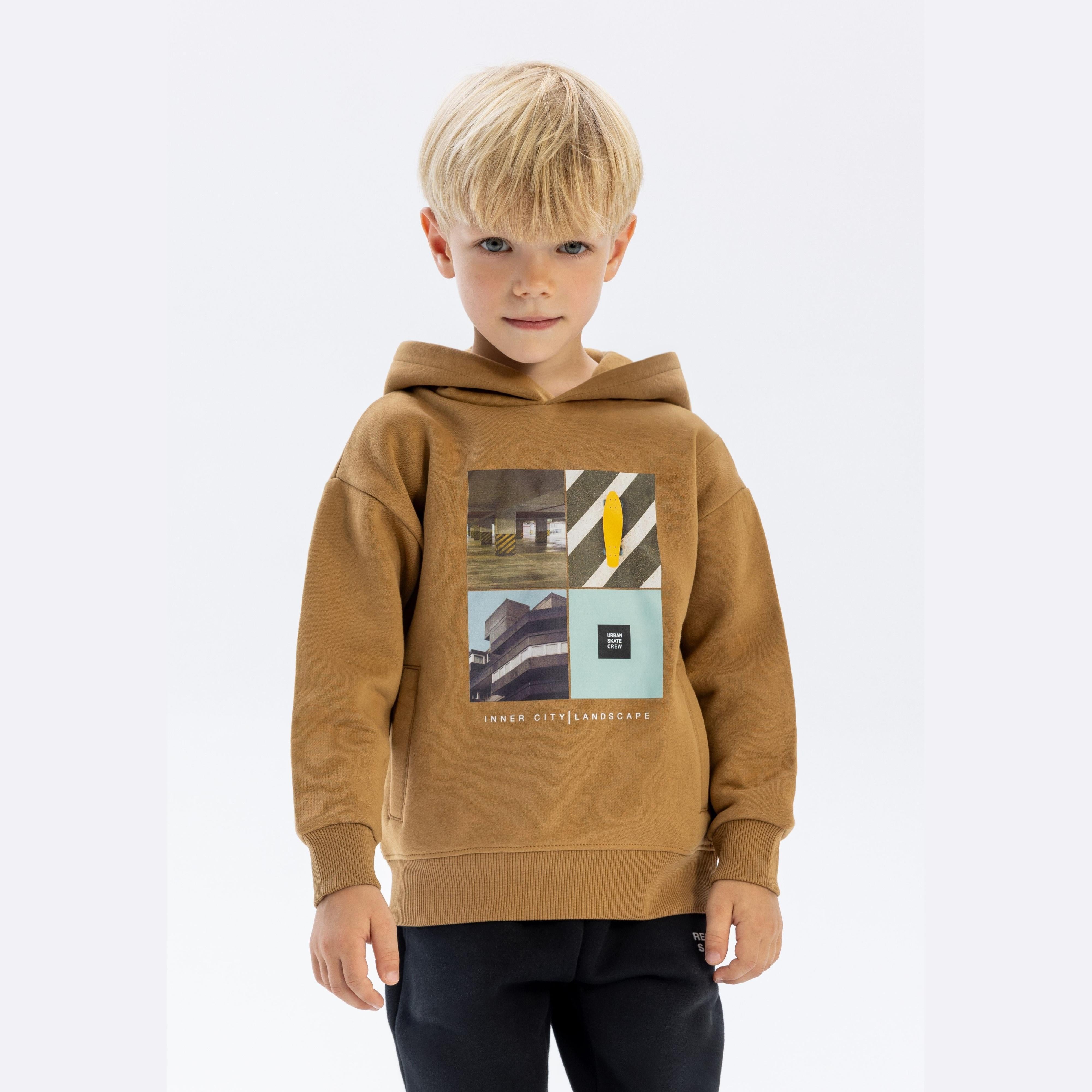 Young blonde boy wearing beige hoodie with urban photographic prints, standing against white background, casual pose