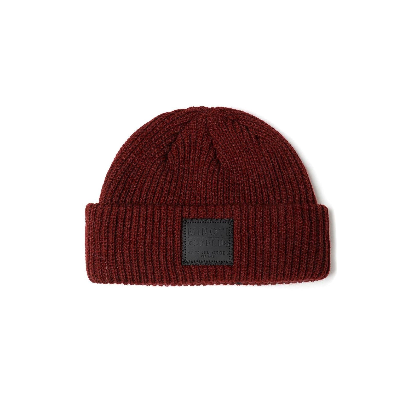 Close-up detail of burgundy ribbed knit beanie showing the embossed dark grey Minoti Surplus patch with Apparel Goods Est. 1967 text and decorative stitching around edges