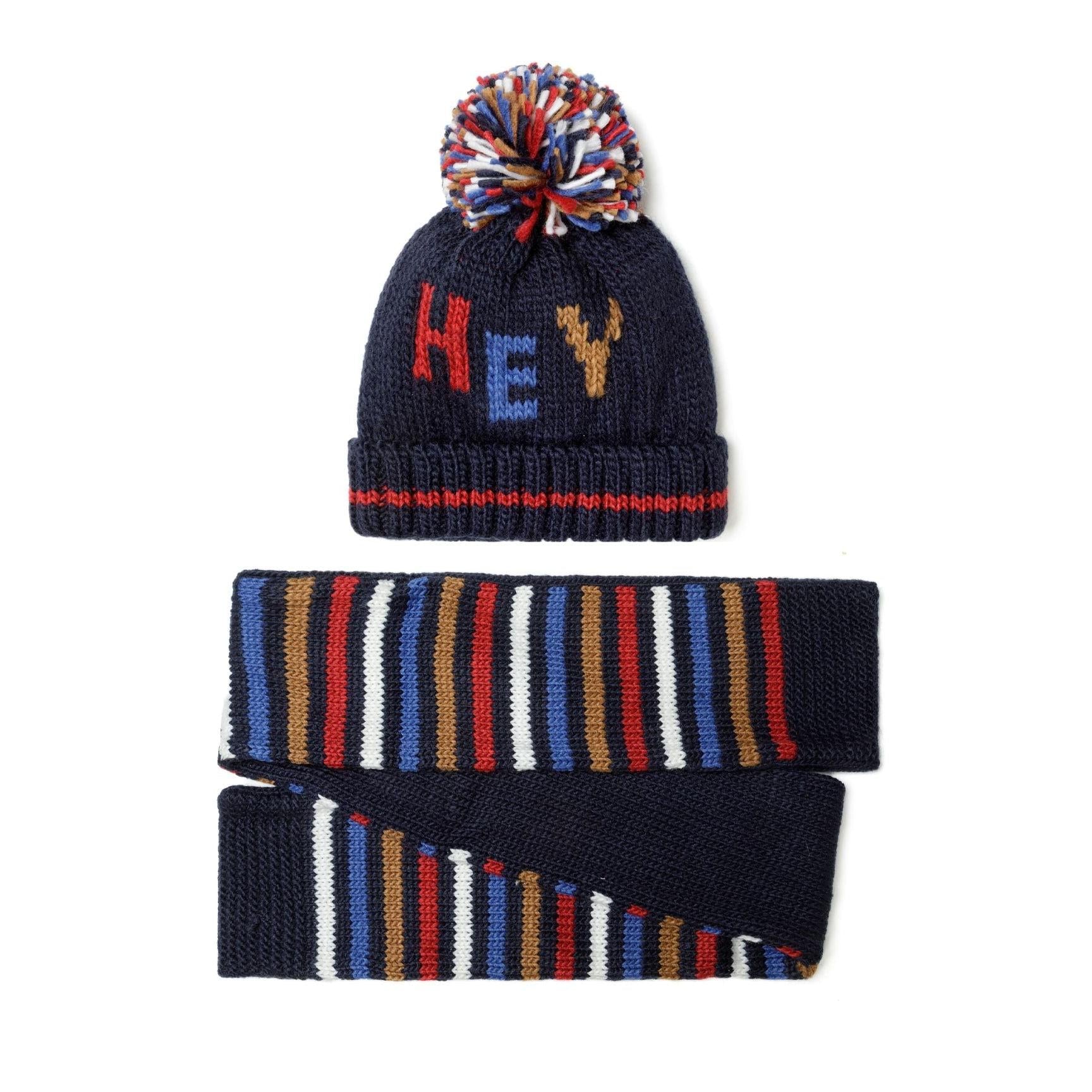 Close-up detail of navy knitted hat showing brown stag motif and multicoloured pom pom against white background
