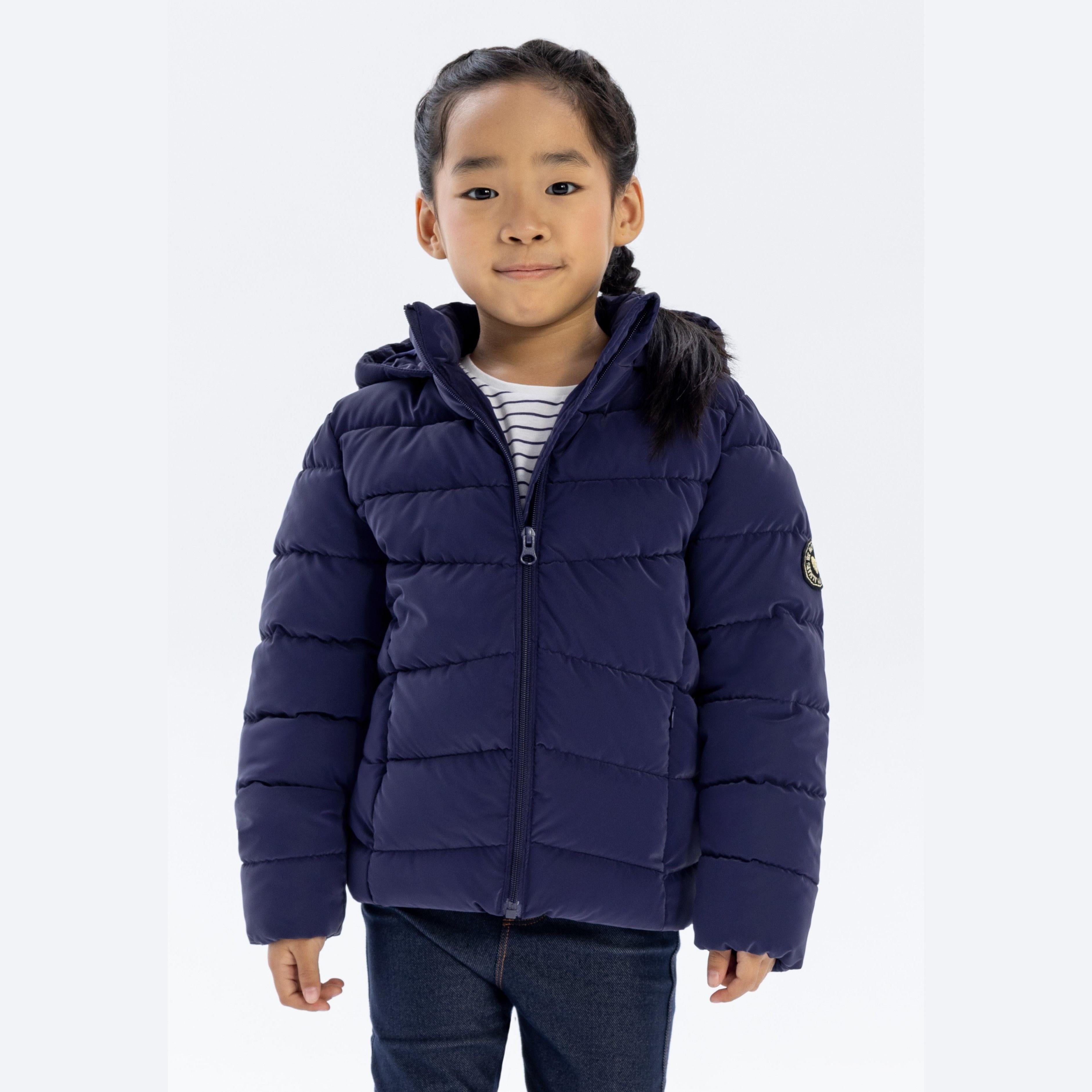 Young girl wearing navy quilted padded jacket with hood and faux fur trim, paired with striped top and jeans, standing against white background