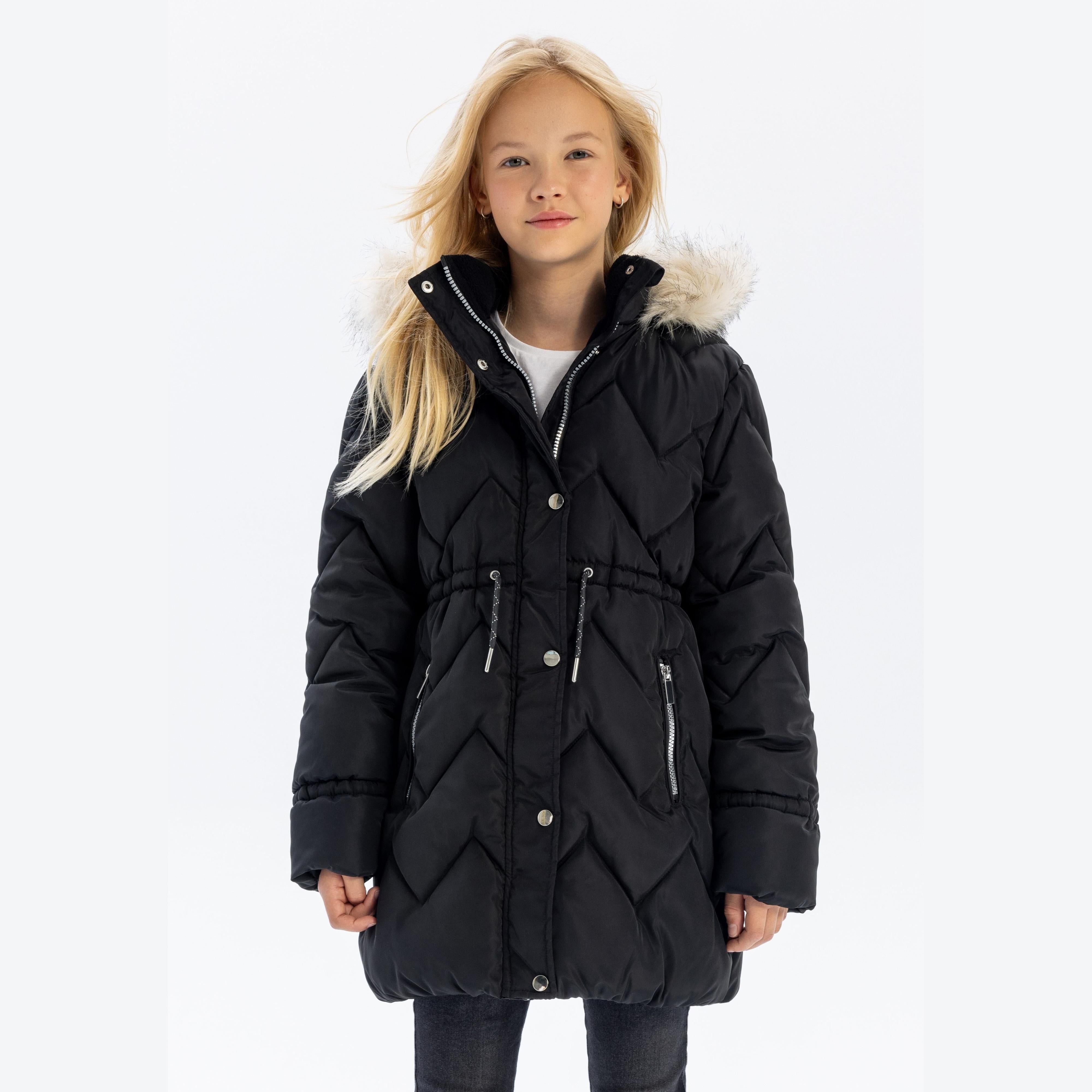 Young blonde girl wearing black quilted parka jacket with faux fur hood, standing against white background, front view showing full length styling