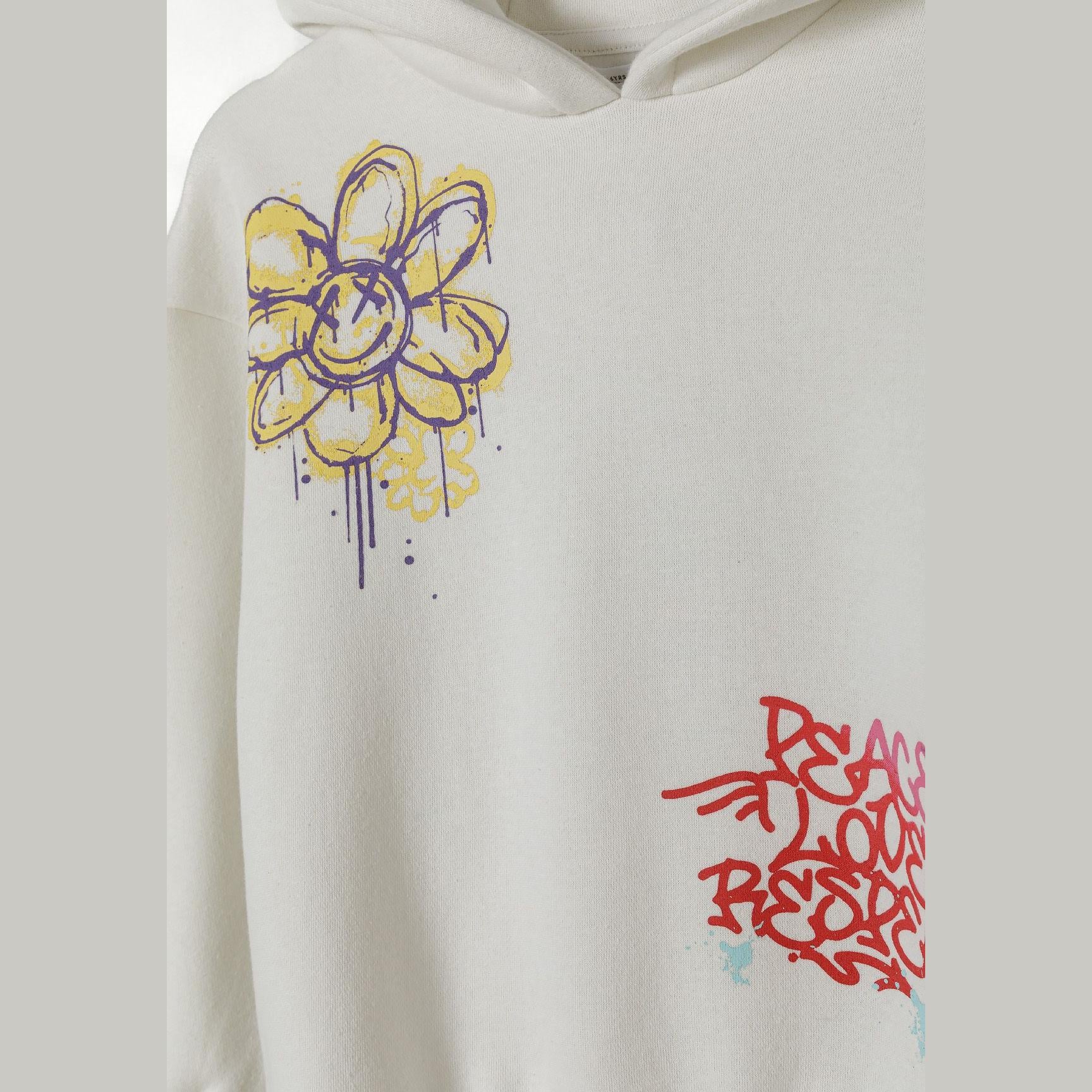 Close-up detail of cream hooded sweatshirt showing yellow and purple hand-drawn flower print and red graffiti text on chest area