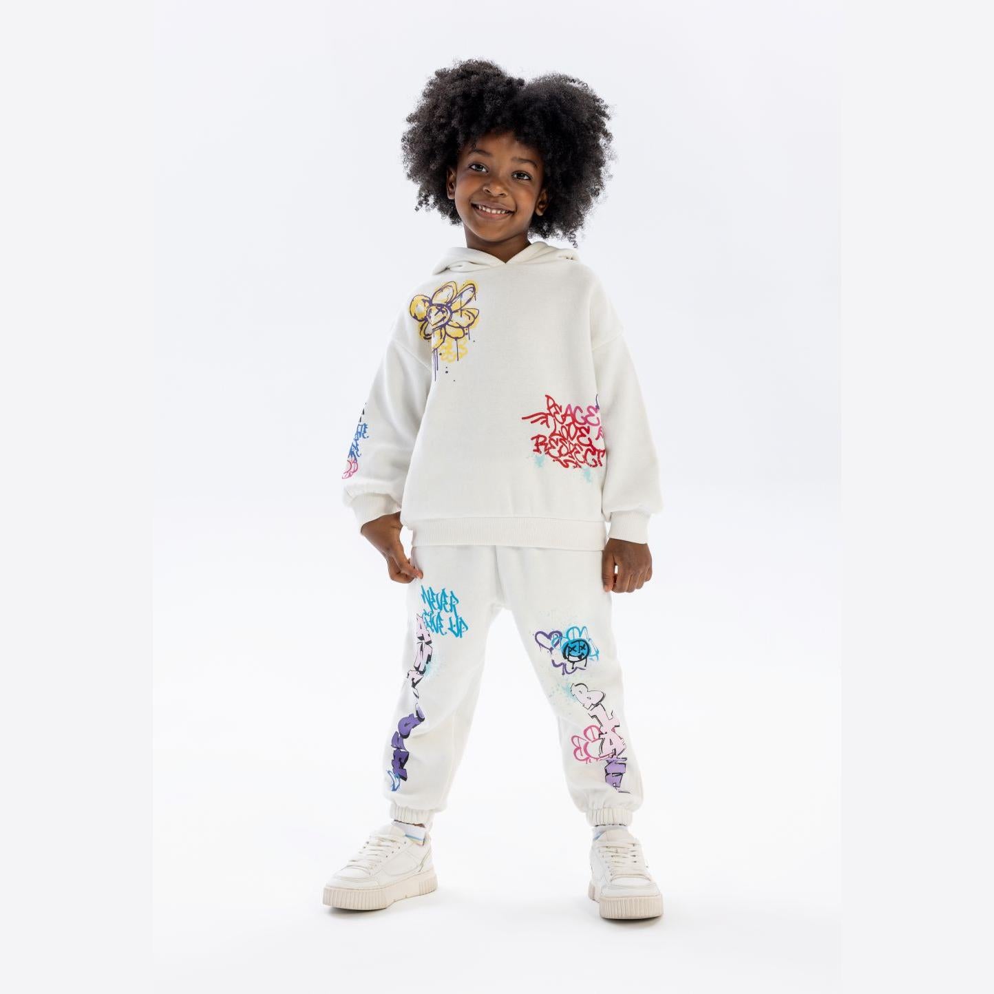 Young girl wearing cream hooded tracksuit with colourful graffiti prints, standing against white background with natural curly hair and white trainers