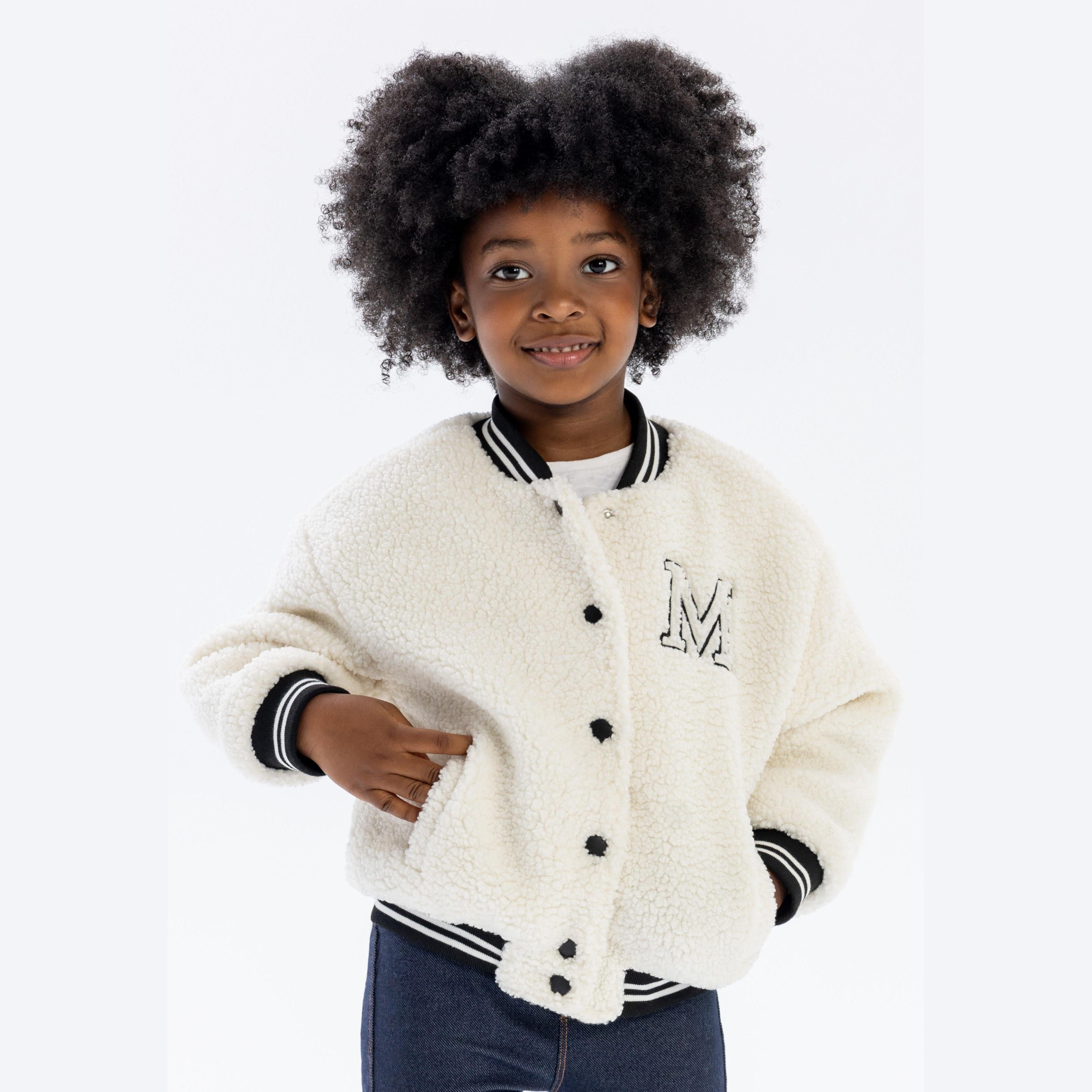 Young girl wearing cream borg baseball jacket with embroidered M detail, paired with jeans, smiling at camera against white background