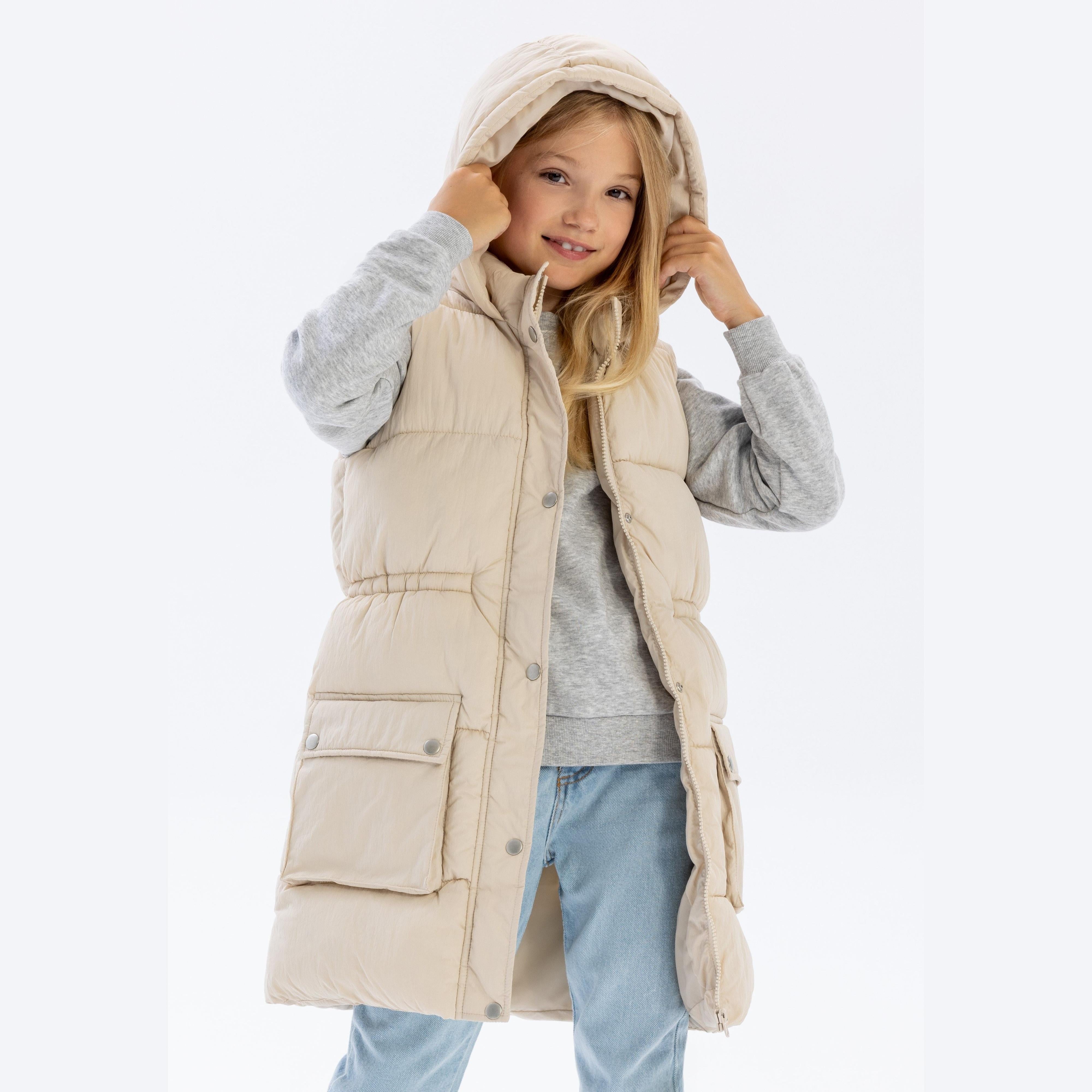 Young blonde girl wearing cream long padded gilet with hood over grey hoodie and jeans, smiling while adjusting hood against white background