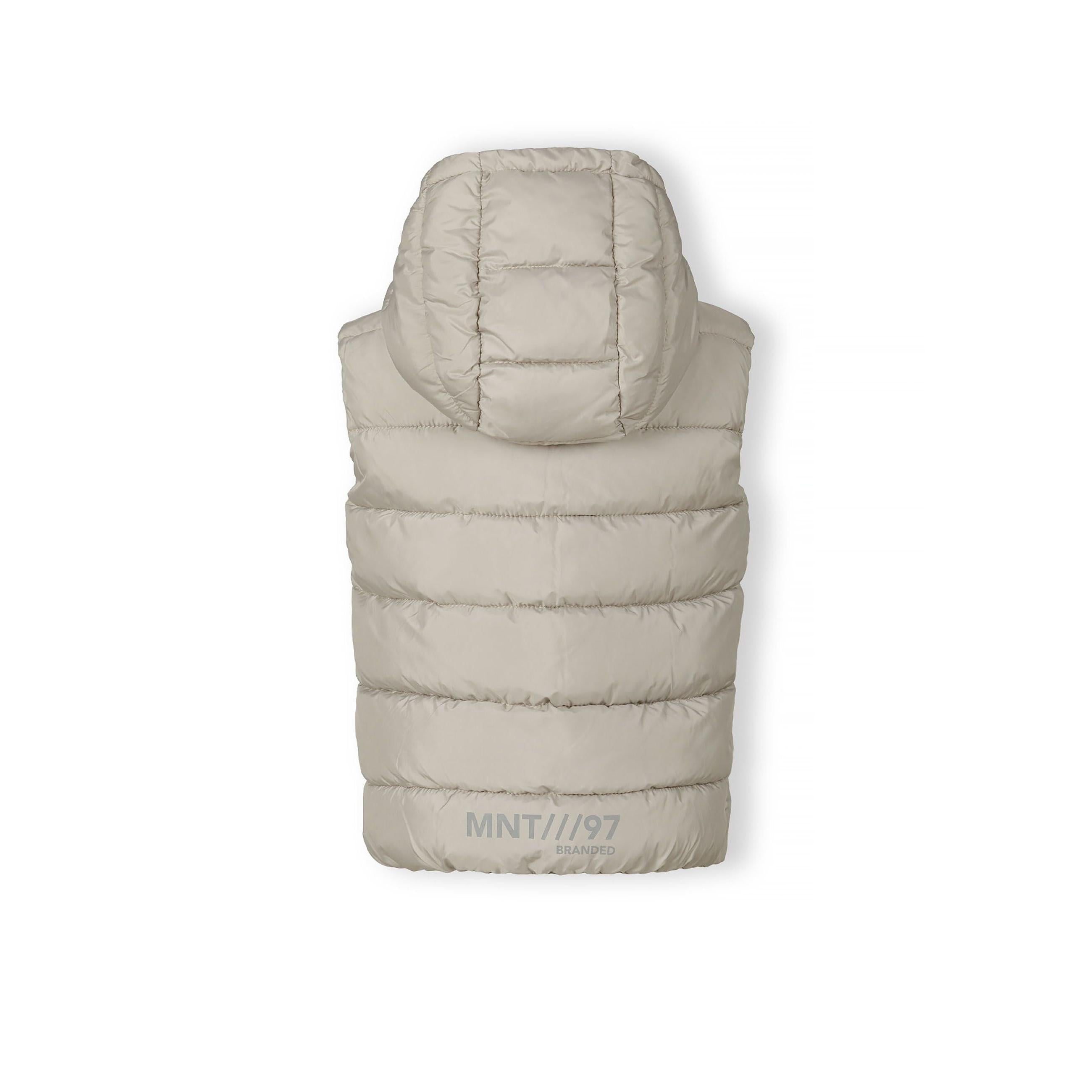 Close-up detail of grey padded gilet showing black circular Minoti logo badge and black zip with reflective zipper pull