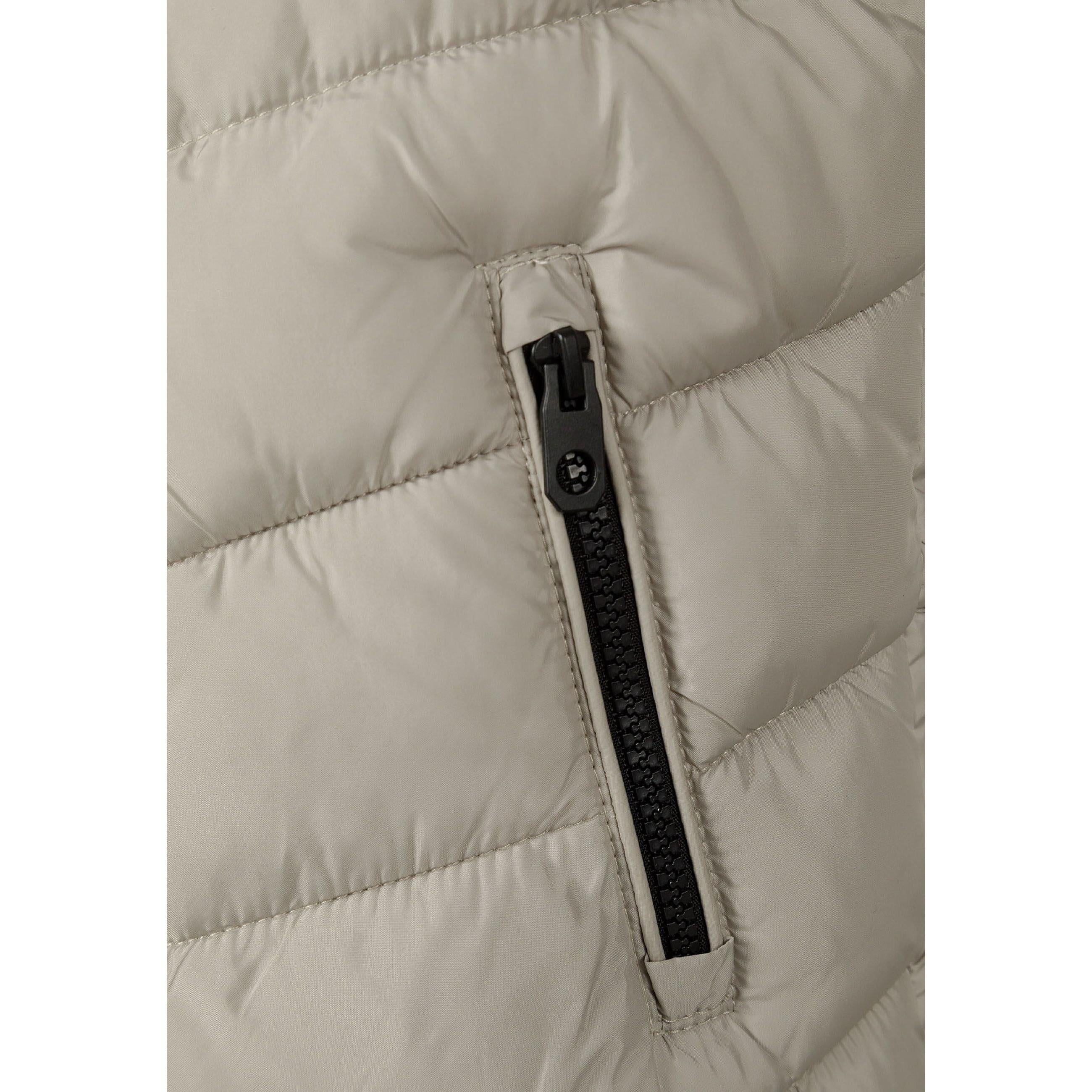 Close-up detail of grey padded gilet showing black circular Minoti logo badge and black zip with reflective zipper pull