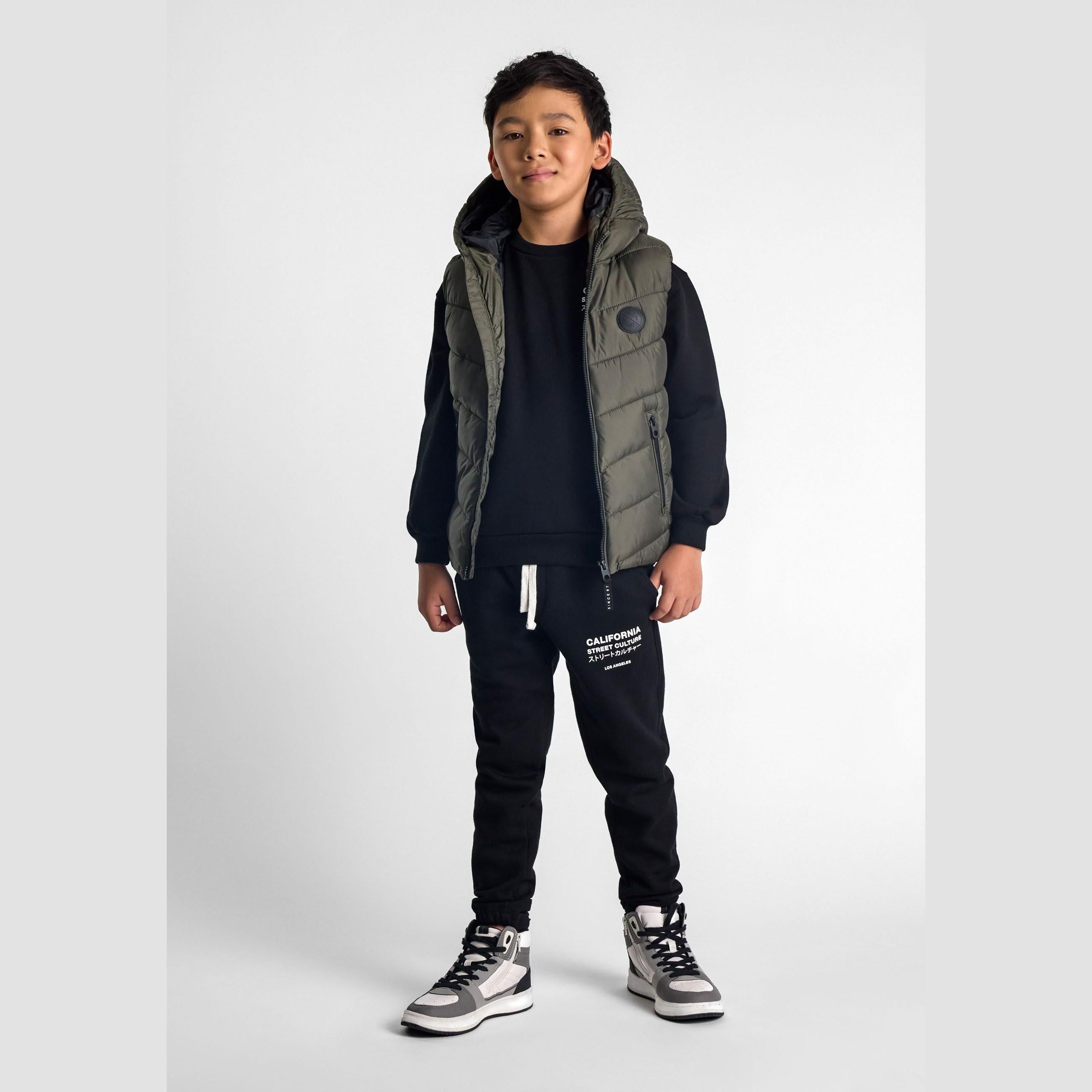 Young boy wearing khaki padded gilet with hood over navy tracksuit against white background, full body front view
