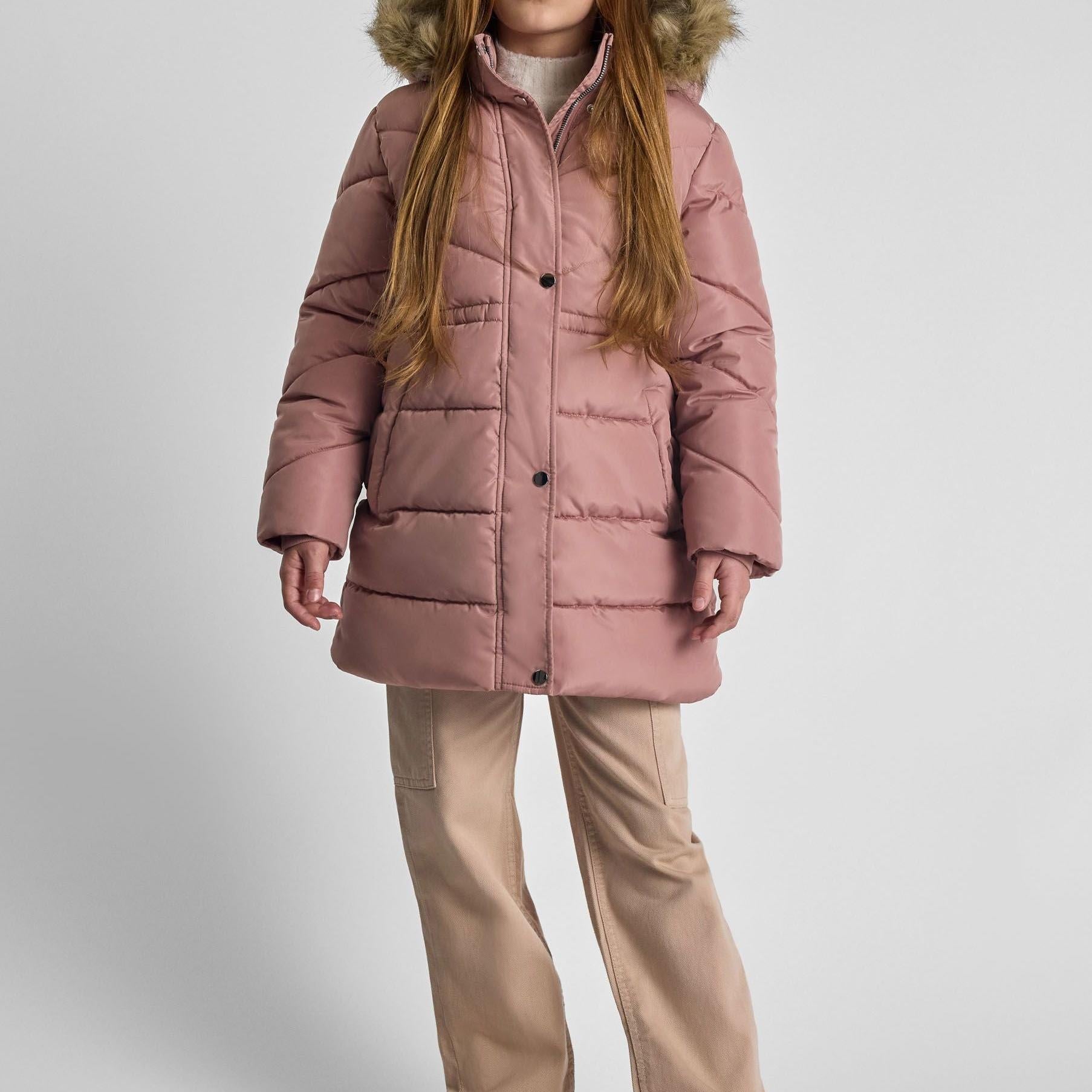 Girl wearing dusty pink quilted winter coat with faux fur hood trim, standing against white background wearing beige trousers
