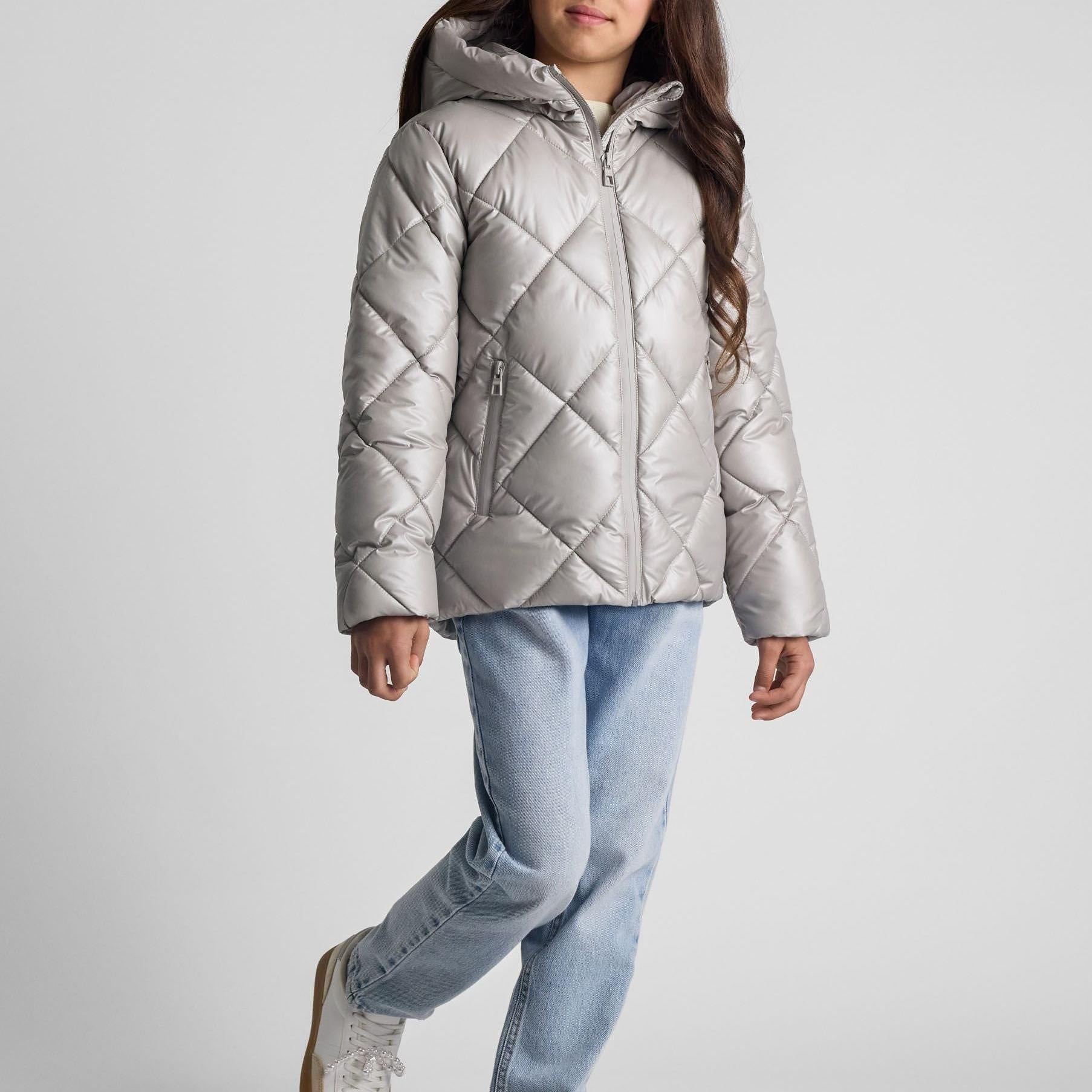 Young girl wearing silver quilted jacket with hood and high collar, paired with light blue jeans, standing against white studio background