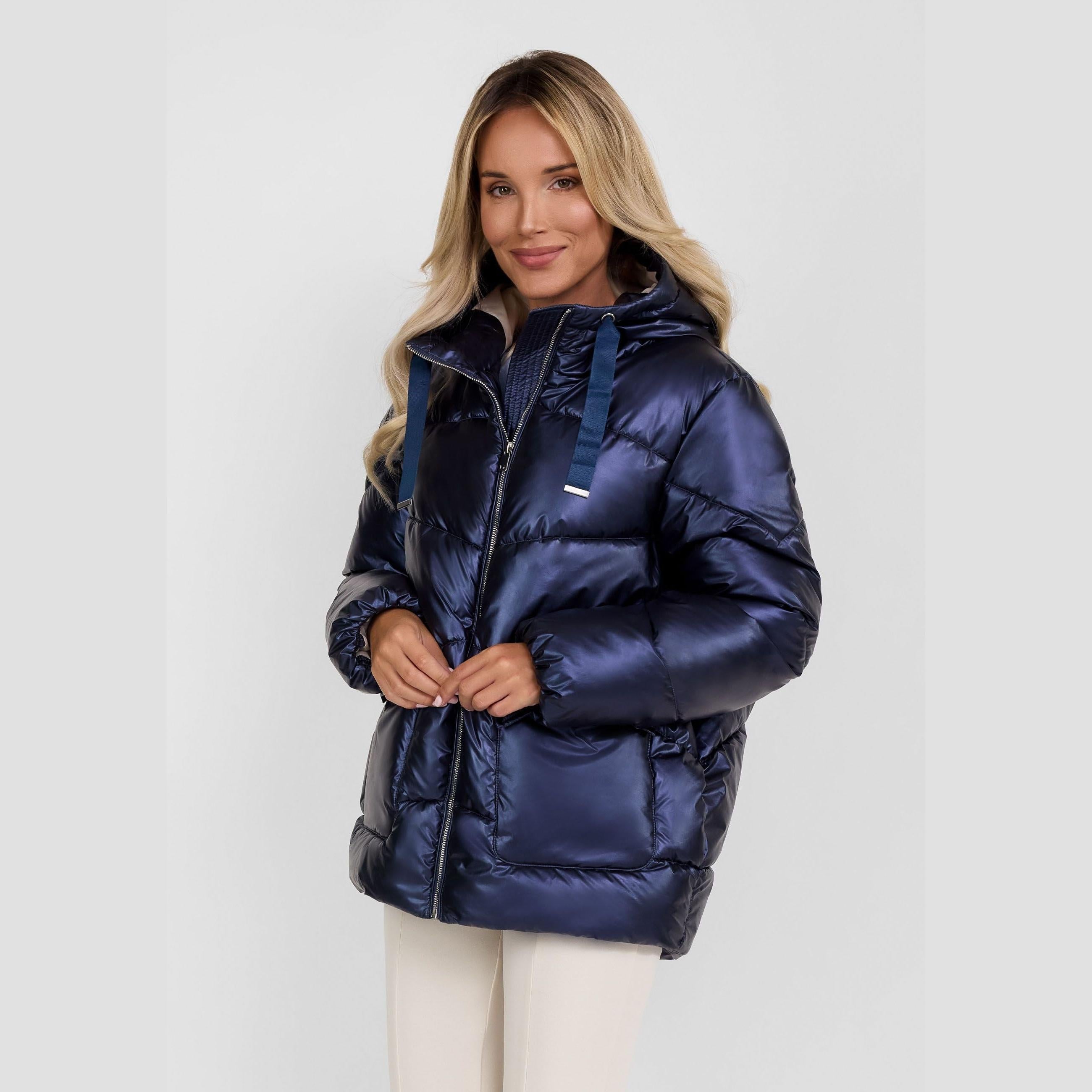 Woman wearing navy quilted puffer jacket with hood up, zipped closure and cream trousers against white studio background