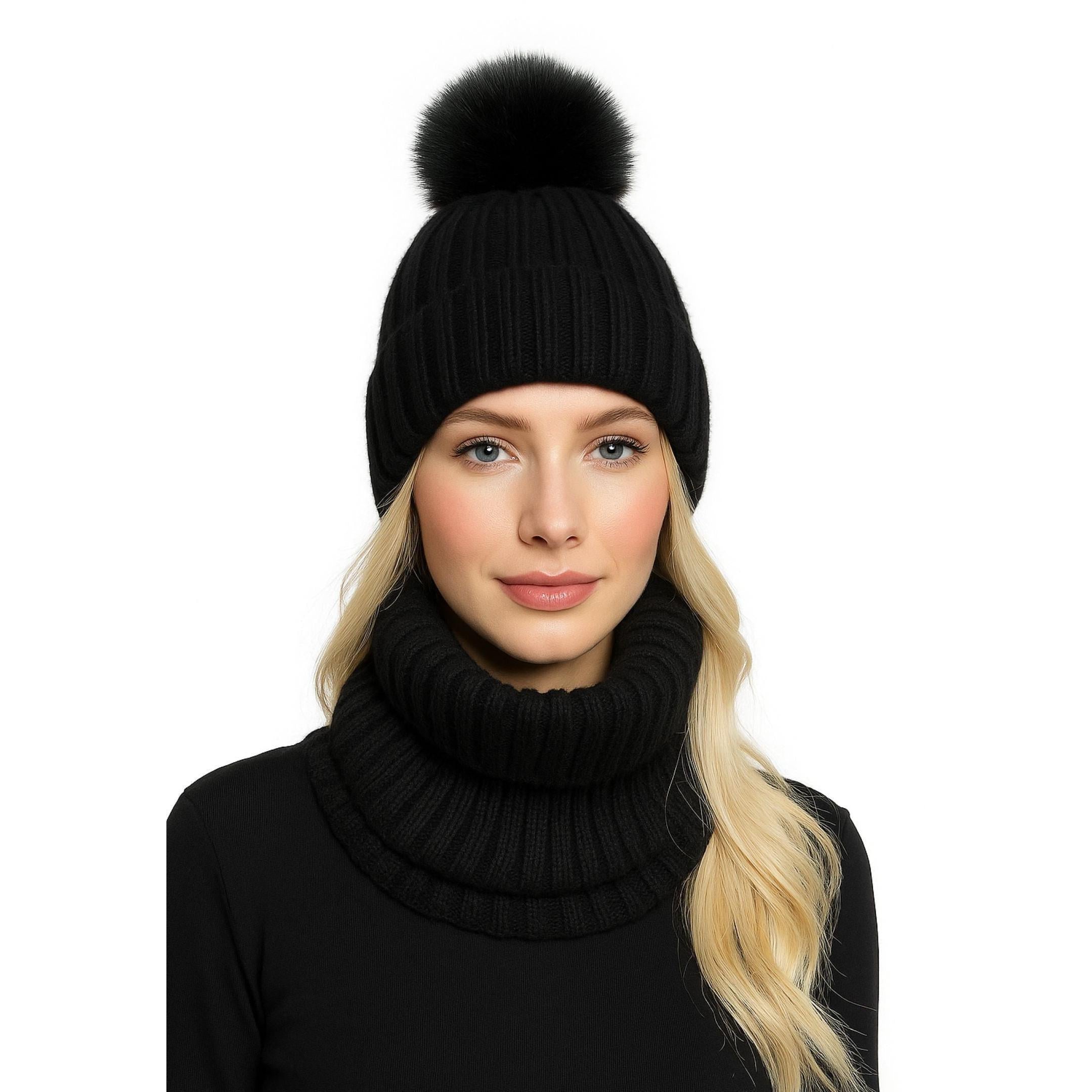 Young blonde woman wearing black ribbed knitted beanie hat with faux fur pom pom and matching black ribbed knit snood, styled with black turtleneck sweater against white background