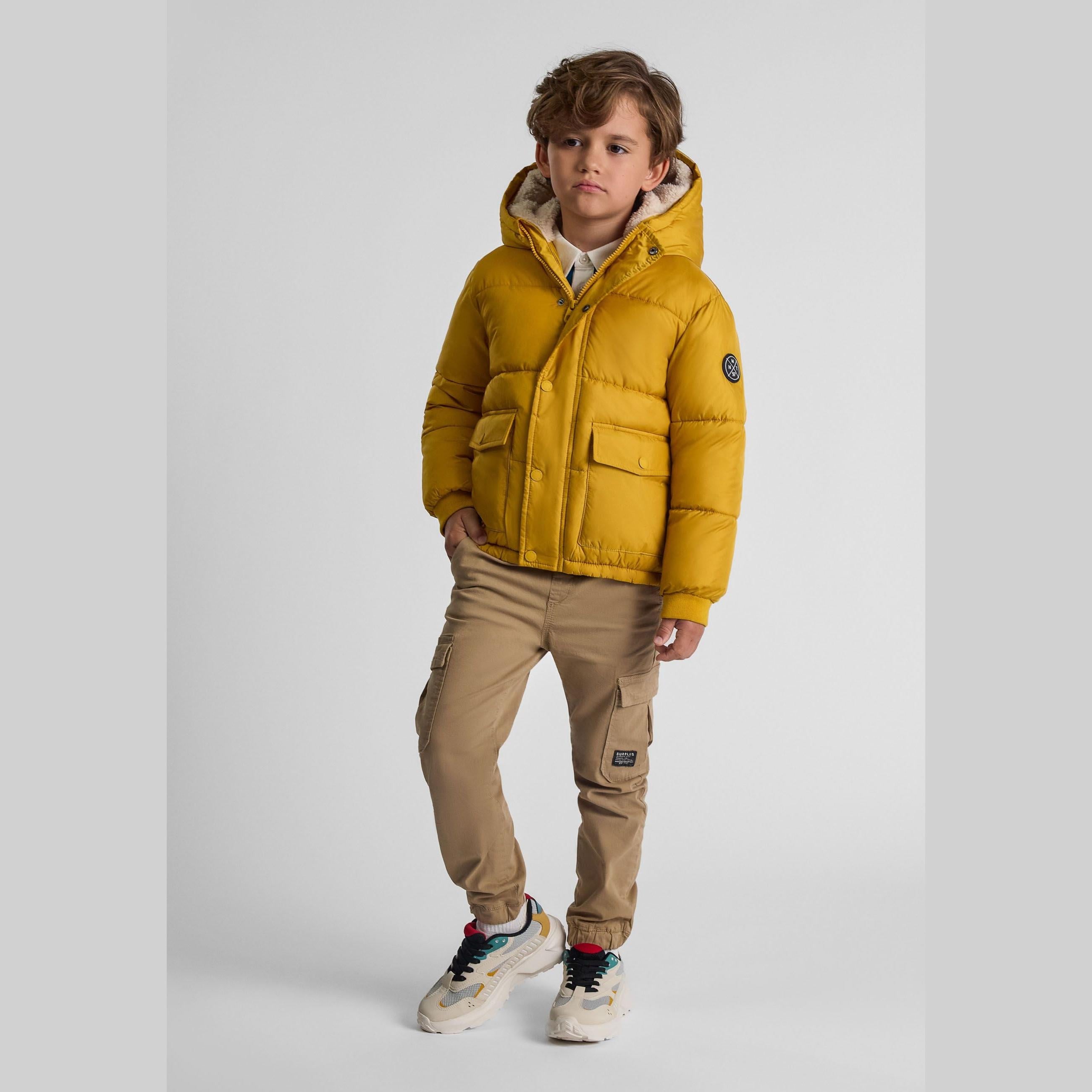 Boy wearing mustard yellow teddy lined hooded jacket with beige cargo pants and sneakers, standing against light grey background