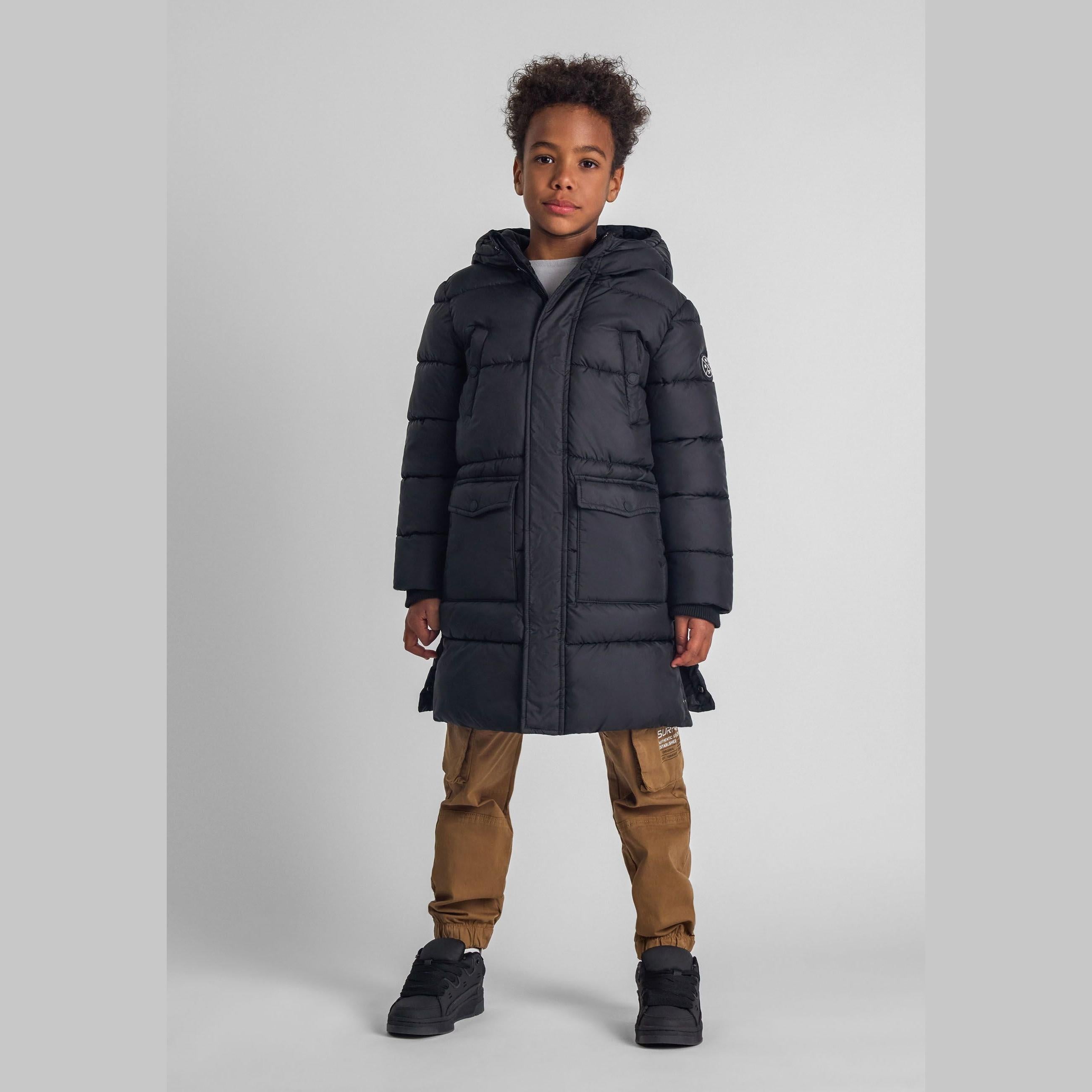 Young boy wearing black hooded long padded coat with brown cargo trousers and black trainers against white studio background