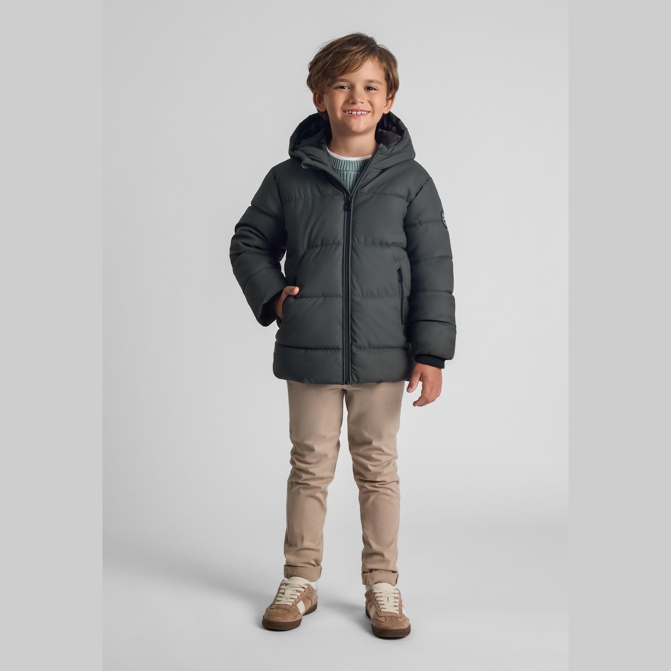 Young boy wearing khaki padded jacket with hood and beige trousers, smiling against white studio background