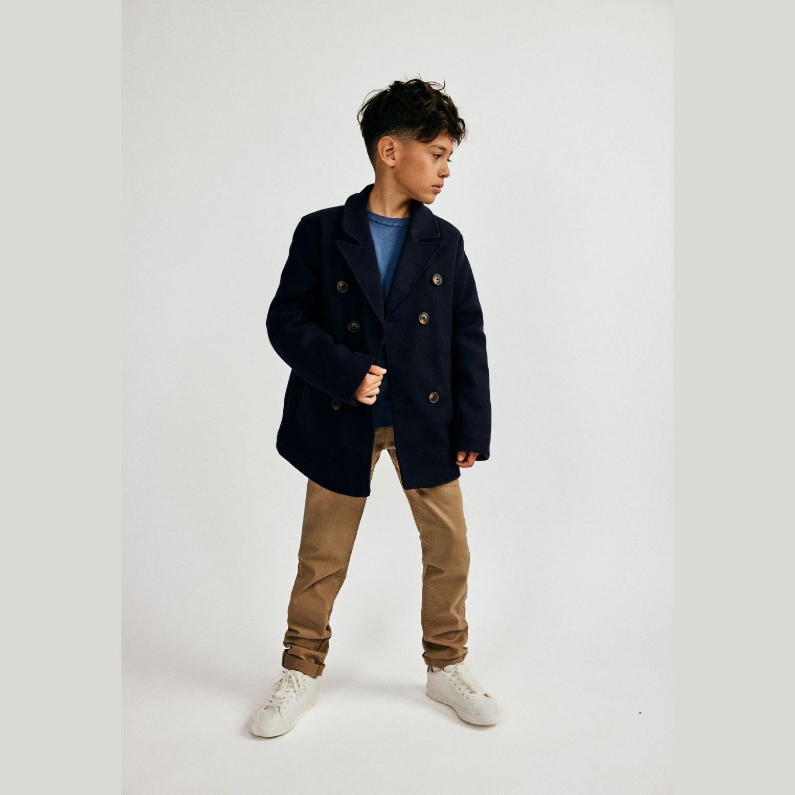 Young boy wearing navy double breasted coat over blue top and beige trousers, standing against white background in studio setting