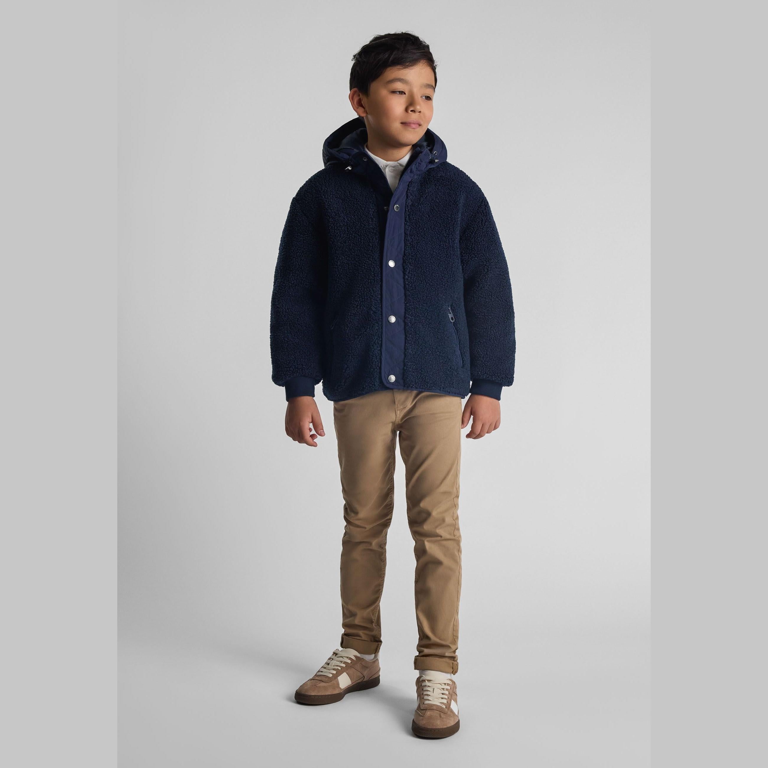 Young boy wearing navy borg jacket with snorkel hood, beige trousers and brown shoes, standing against white background