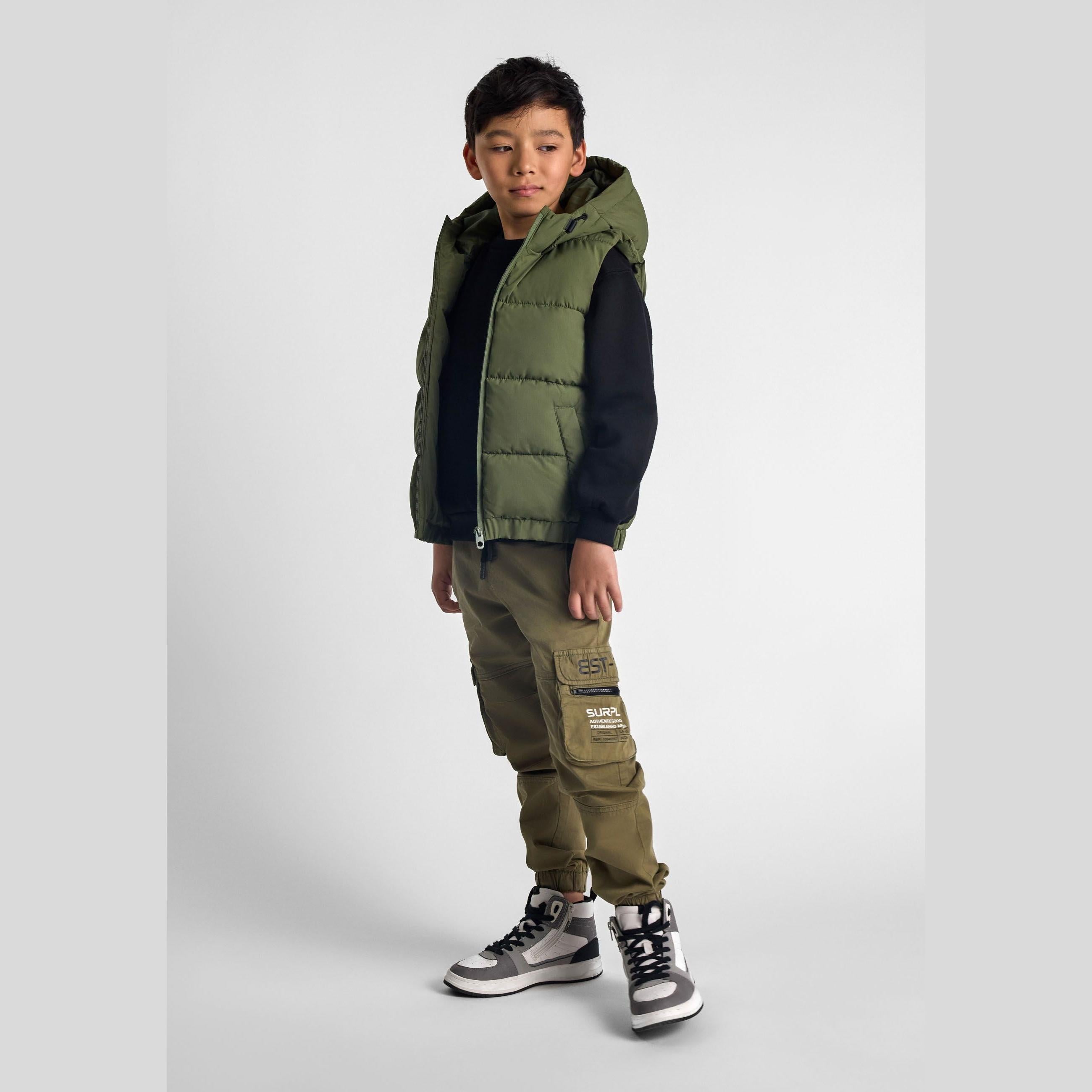 Boy wearing khaki padded gilet with hood over black sweater and matching khaki cargo pants with patches, standing against white background