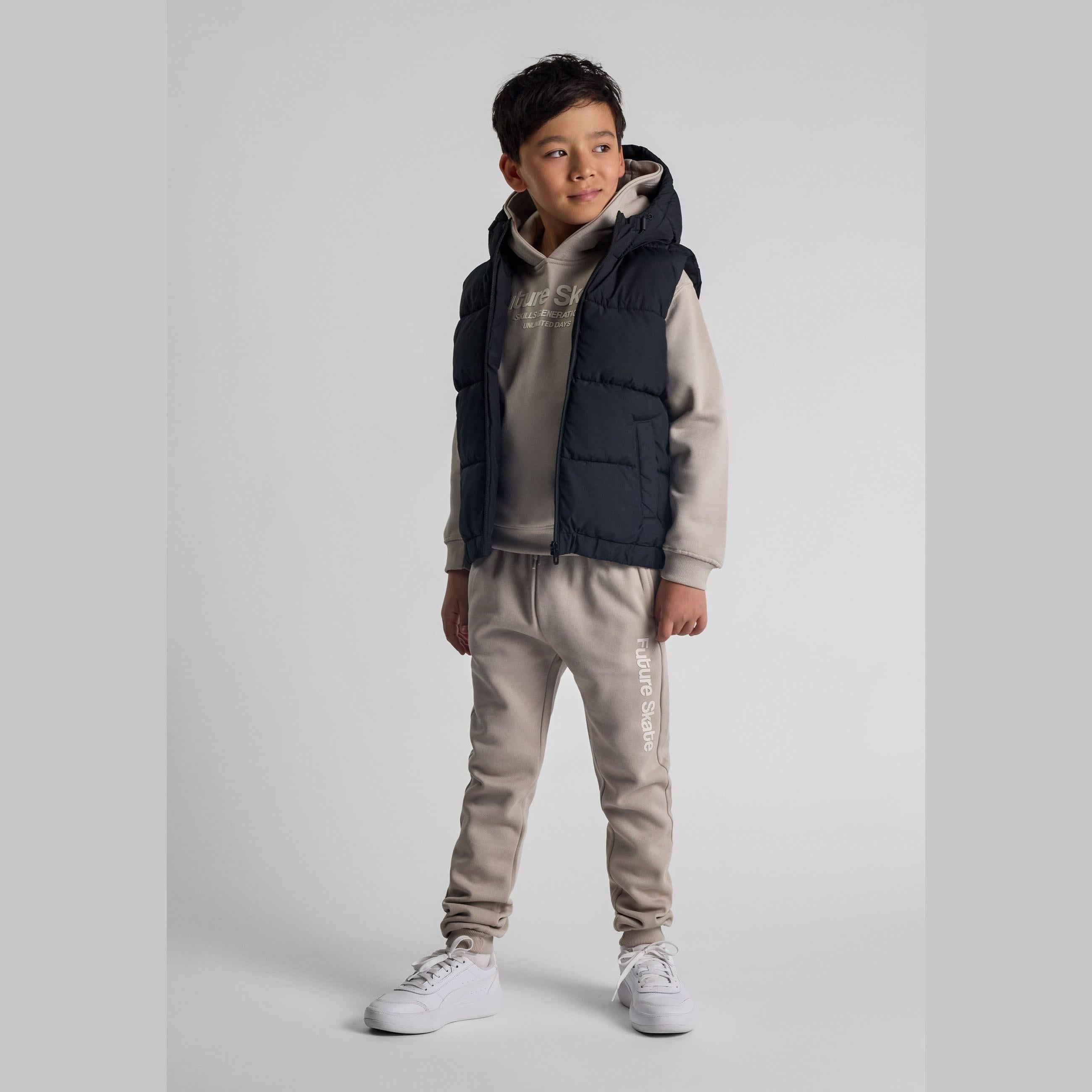 Young boy wearing black hooded padded gilet over grey tracksuit, standing against white studio background with hands in pockets