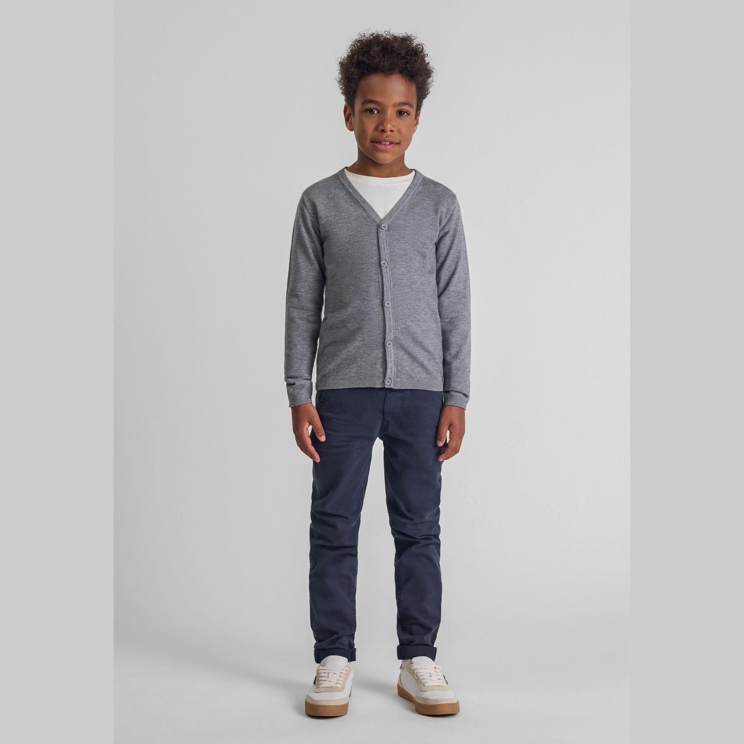 Young boy wearing grey fine knit cardigan with navy trousers and white sneakers against neutral background, front view full body shot