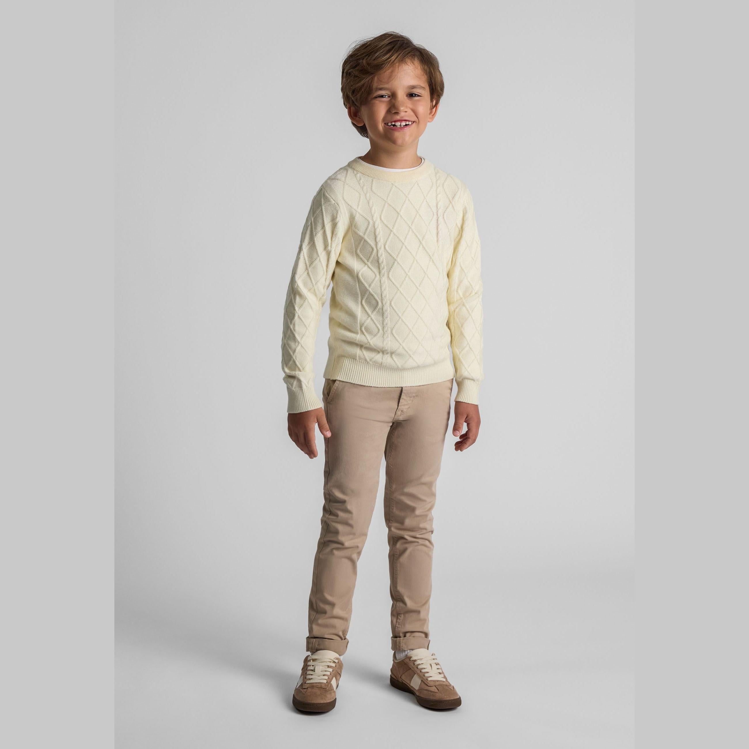 Young boy wearing cream cable knit jumper with beige chinos against grey background, full body studio shot