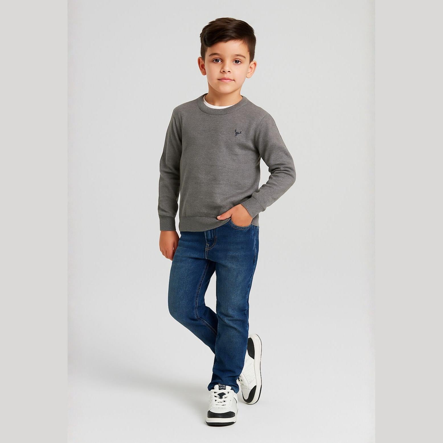 Young boy wearing grey fine knit crew neck jumper with blue jeans and white trainers, standing against white studio background