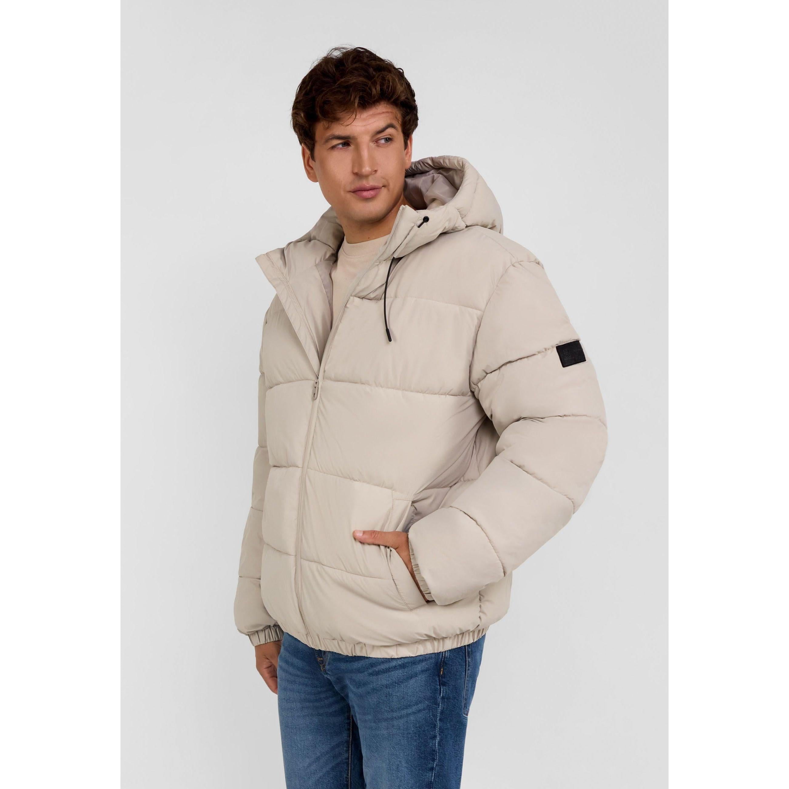 Young man wearing grey ripstop jacket with hood up, standing against light grey background, hands in pockets, paired with blue jeans