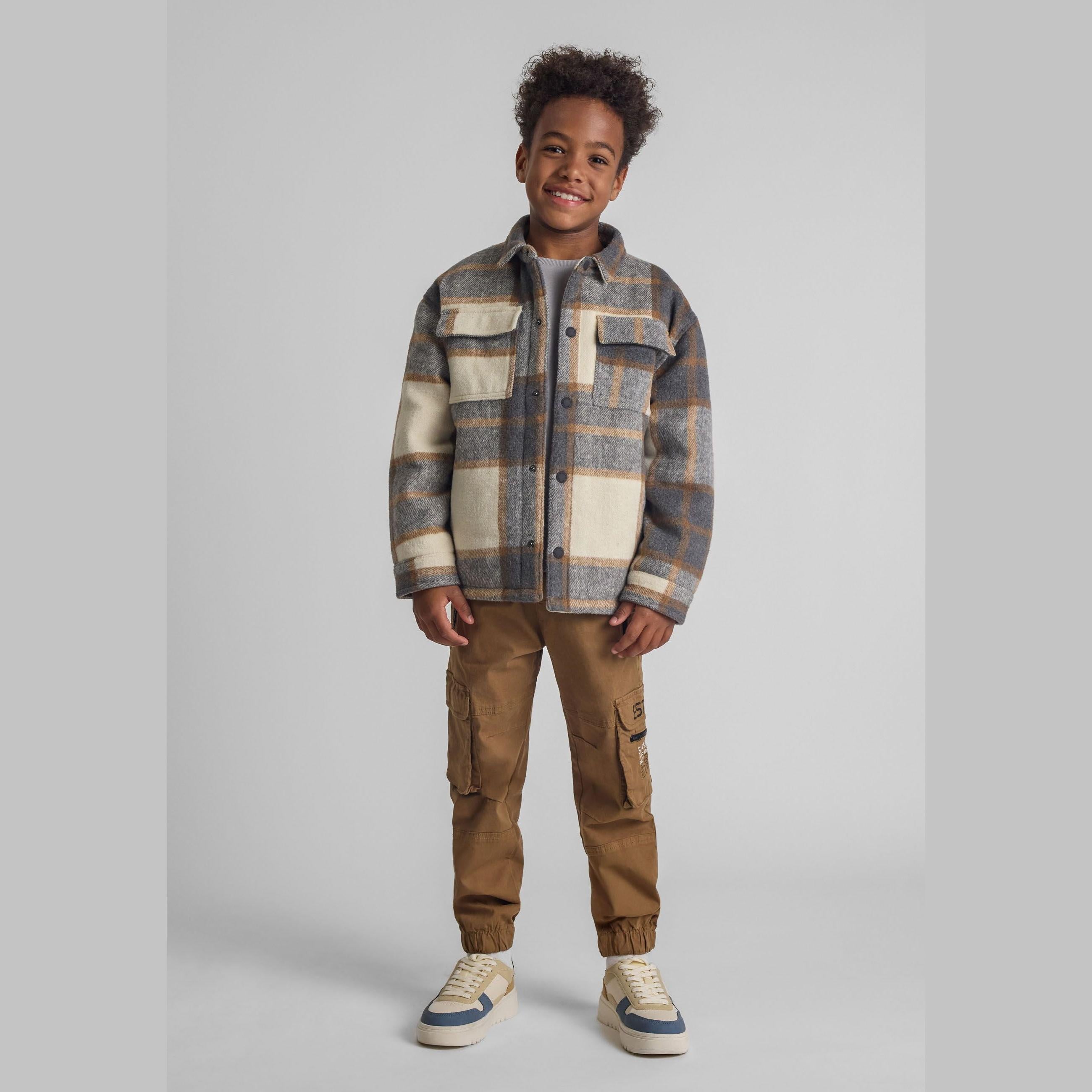 Young boy wearing grey check padded shirt jacket with brown cargo trousers and sneakers, smiling against neutral studio background