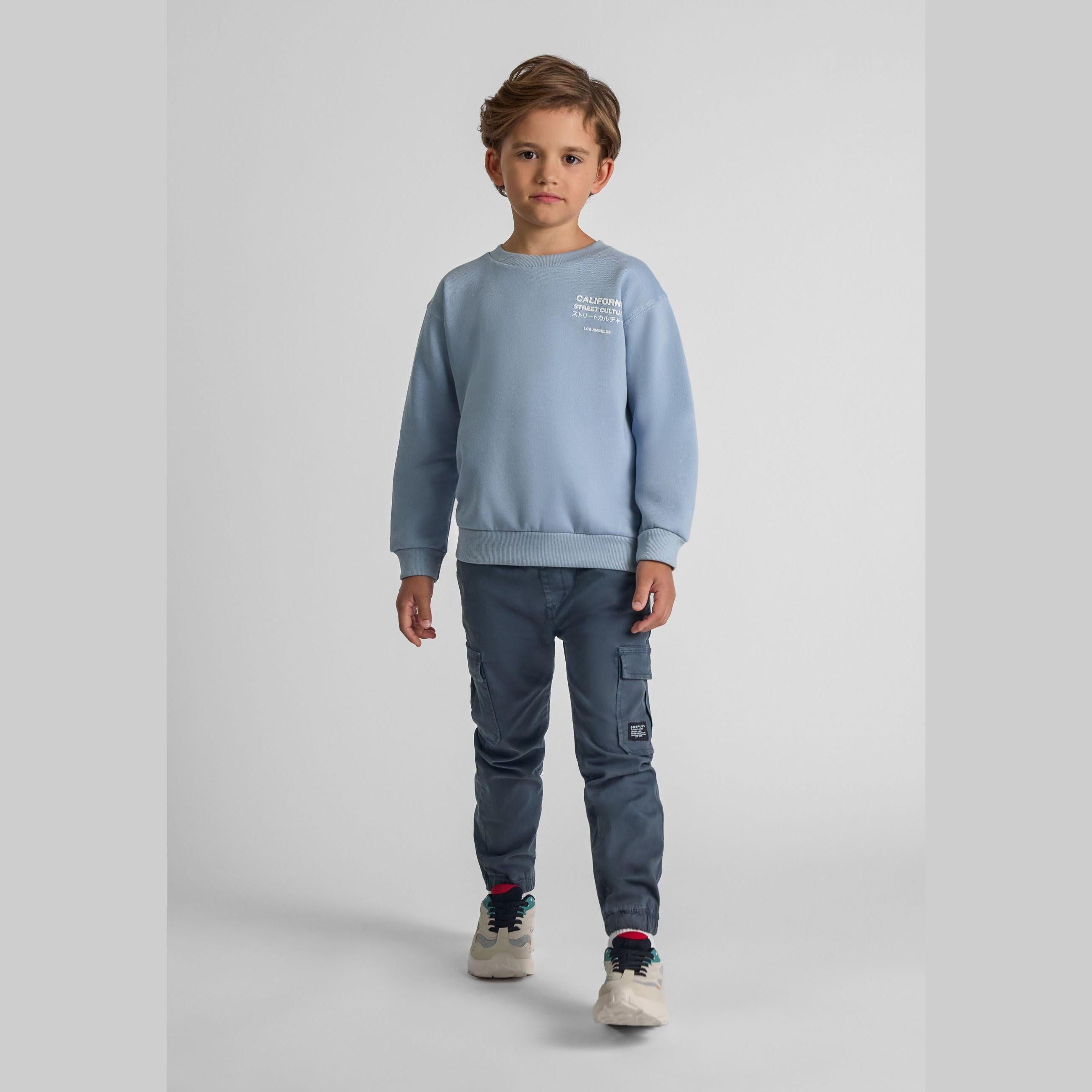 Young boy wearing light blue sweatshirt and navy cargo trousers with elasticated waist, standing against white studio background