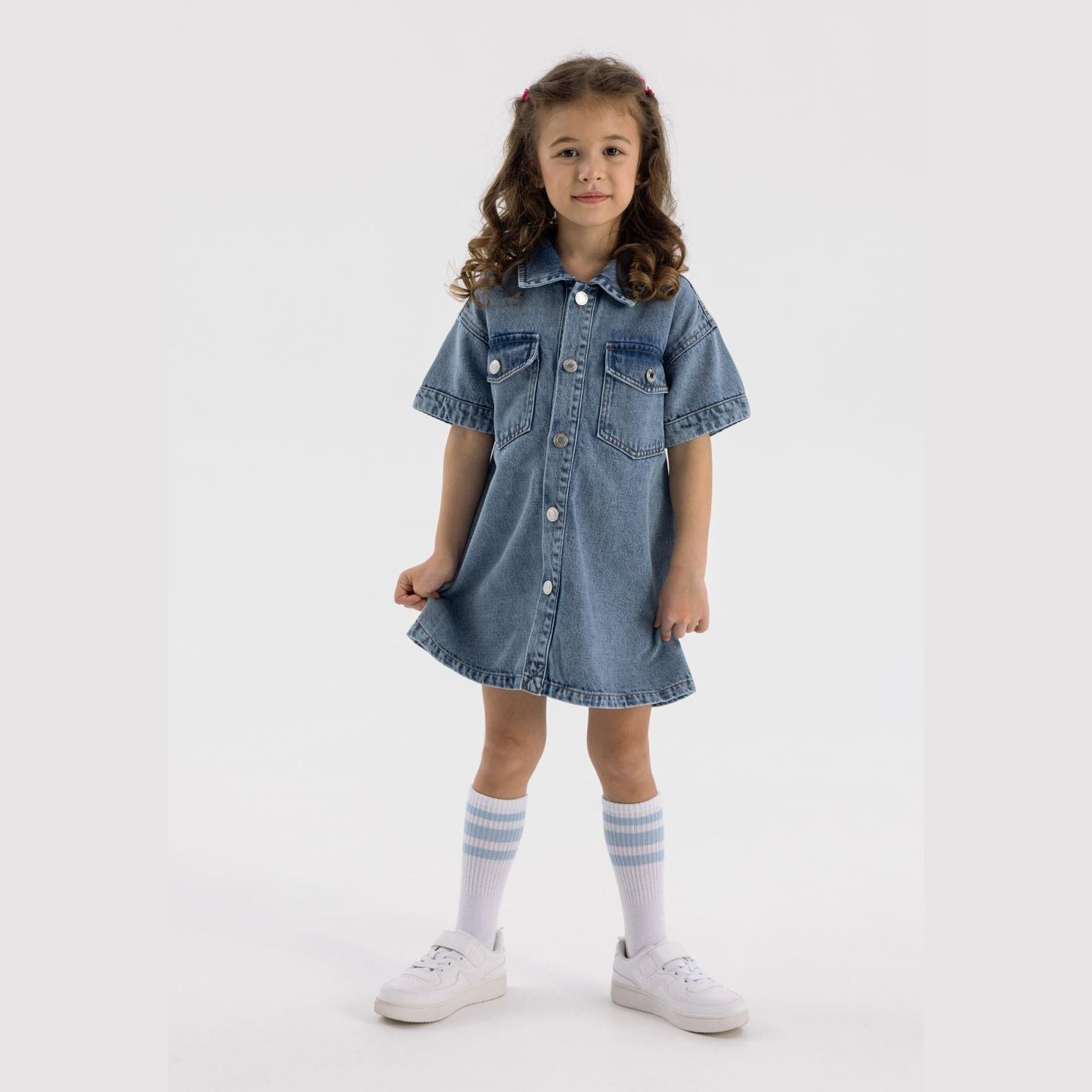 Young girl wearing navy denim shirt dress with short sleeves, collar and button front, styled with striped socks and white trainers against white background