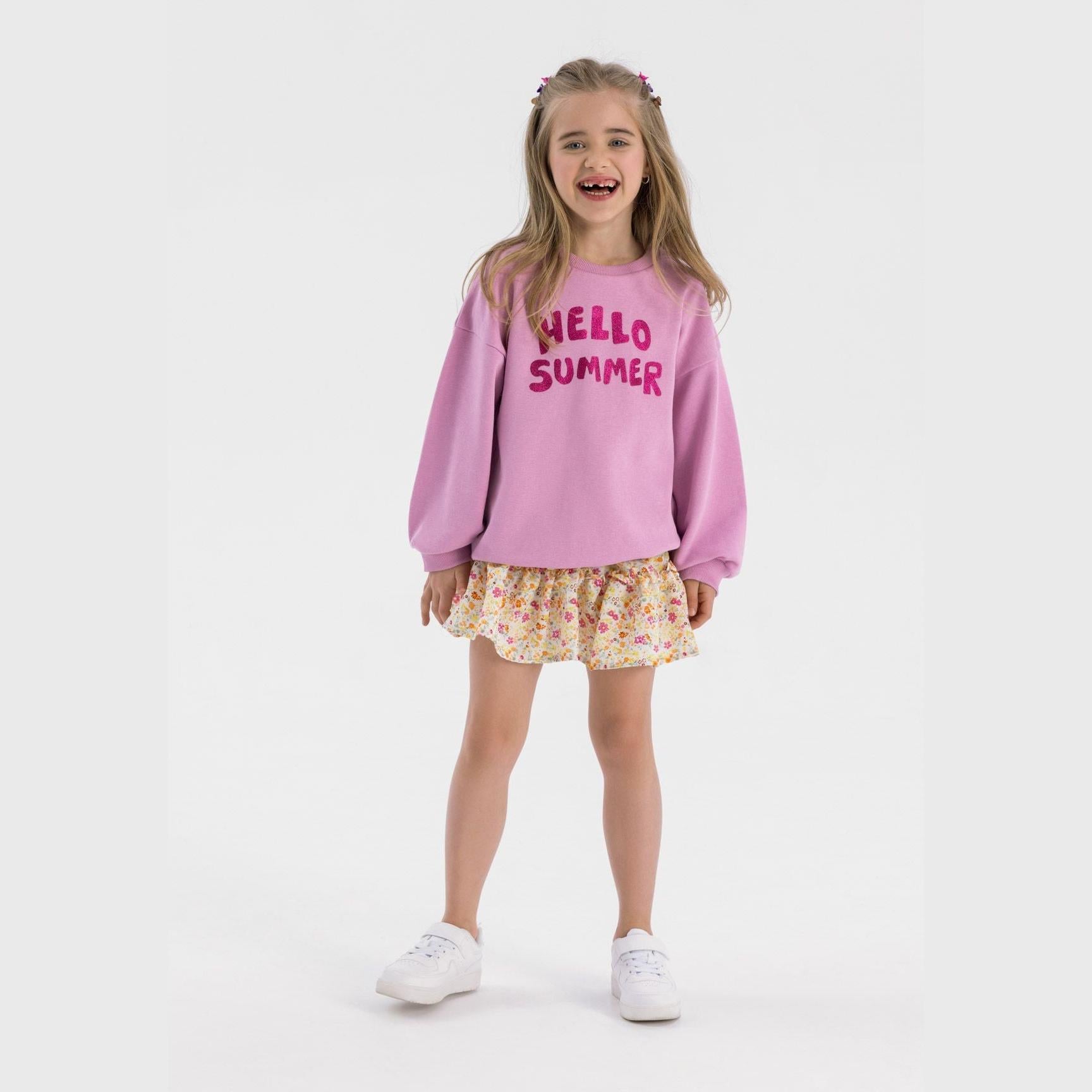 Young girl wearing lilac fleece sweatshirt with Hello Summer print paired with floral skirt, smiling against white background